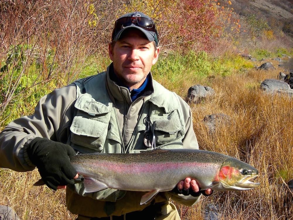 Man holding a large rainbow trout, outdoors by dry grass and shrubs.