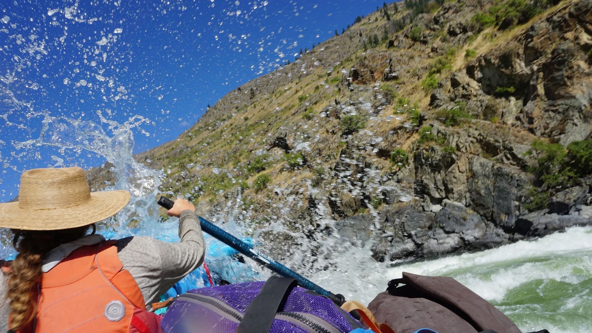 Rafter in a straw hat paddles through whitewater, splashing water. Mountain backdrop, blue sky.