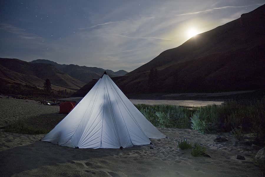 A tent is lit up at night on the shore of a lake.