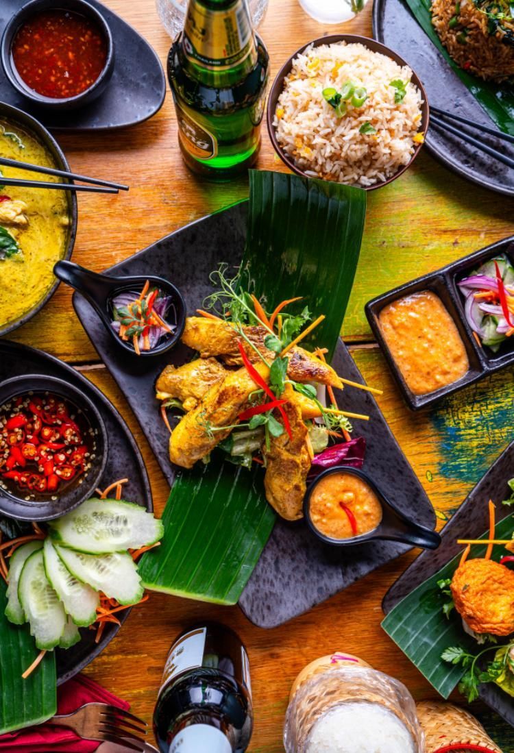 Assorted colorful Asian dishes on banana leaves with rice, sauces, and drinks on a wooden table