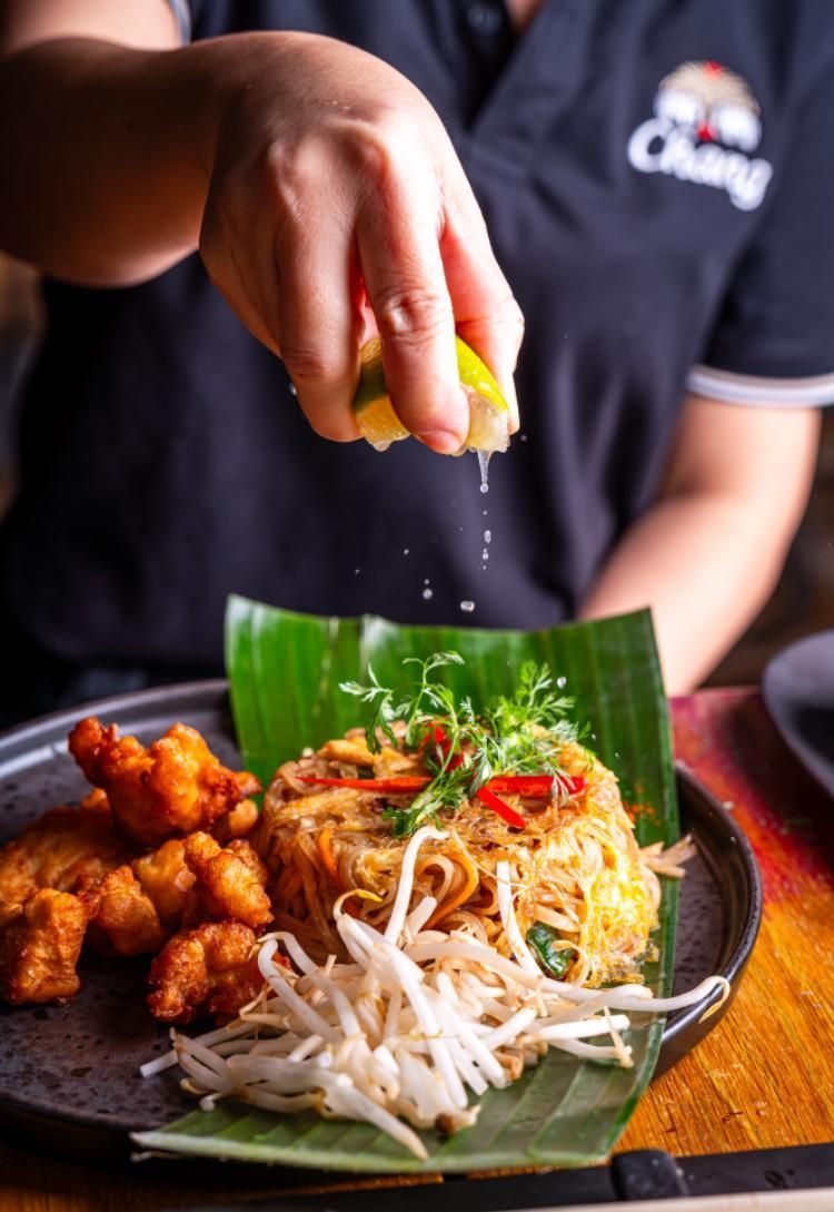 Hand squeezing lime over a plate of noodles, bean sprouts, and fried chicken on a banana leaf plate