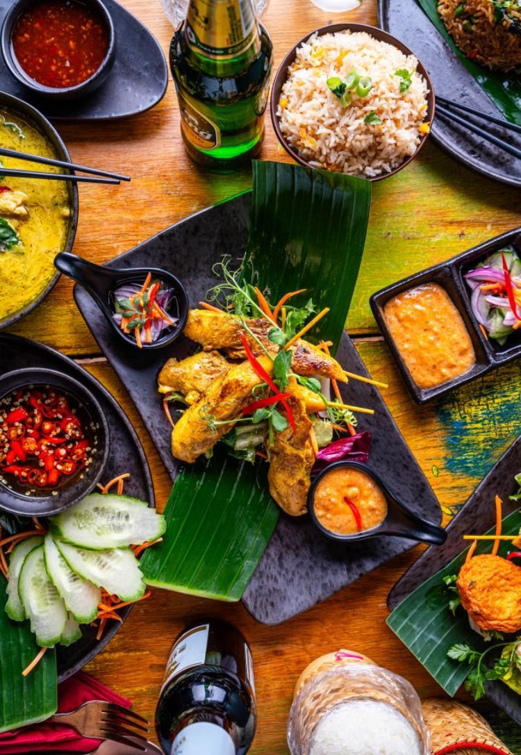 Assorted colorful dishes on banana leaves and black plates, with rice and drinks on a rustic table.