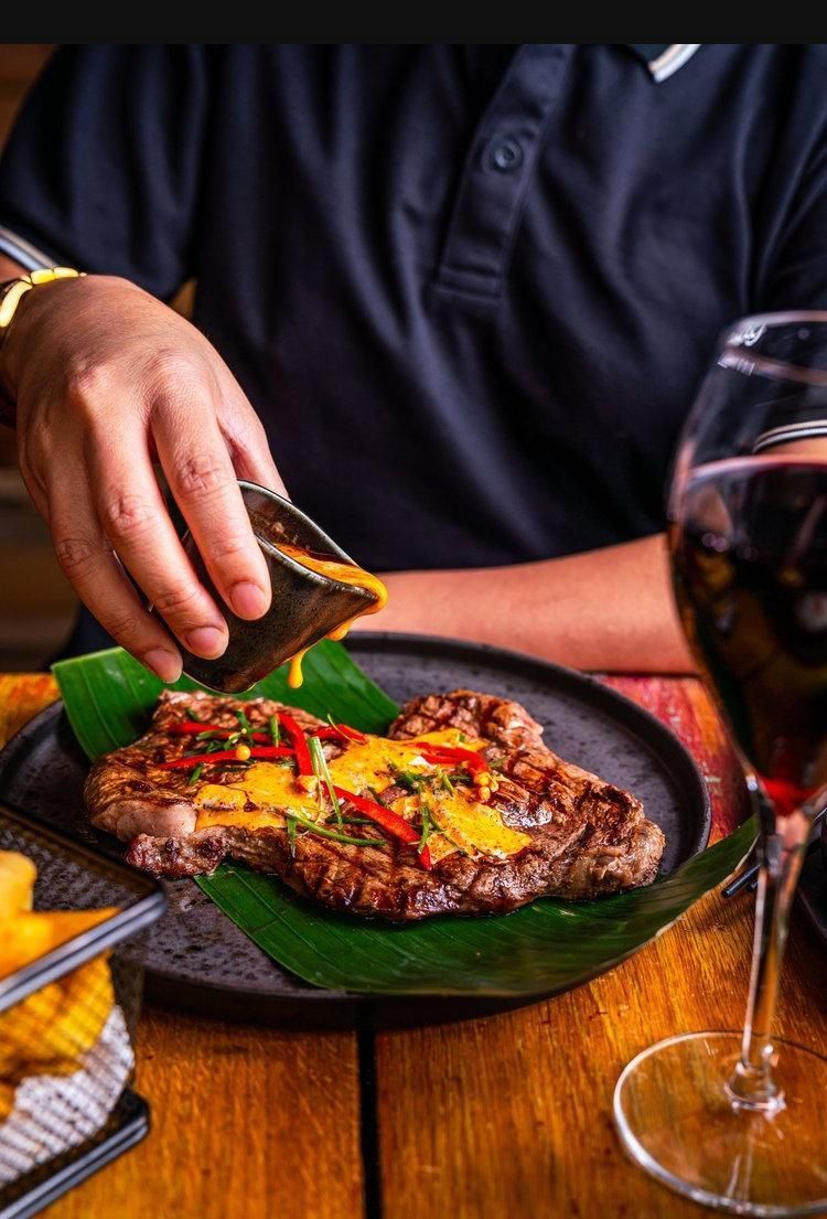 Hand placing a folded taco on a wooden table beside a wine glass and plate of grilled food