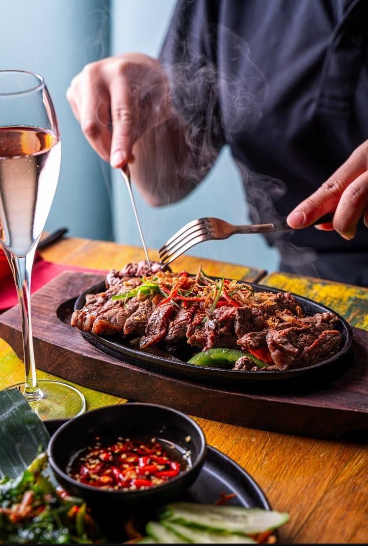 Hands serving sizzling steak on a hot plate with a glass of wine and chili sauce beside it