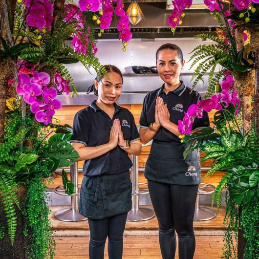 Two restaurant staff in black uniforms stand with hands in prayer pose under pink flowers.