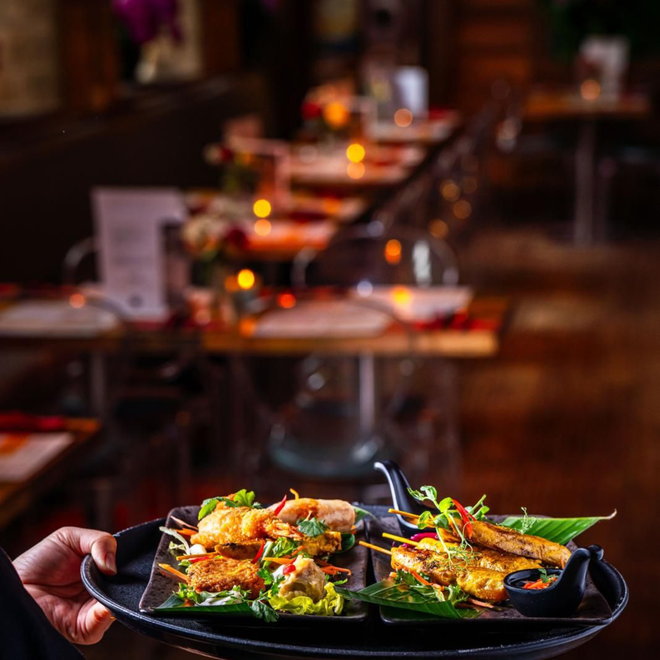 Hand holding a black plate of grilled seafood in a warmly lit restaurant dining room