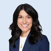 Woman with dark hair smiles, wearing a navy blazer and white top, necklace, against a light blue background.