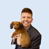 Woman with short hair, wearing a blazer, smiles while holding a long-haired dachshund puppy.