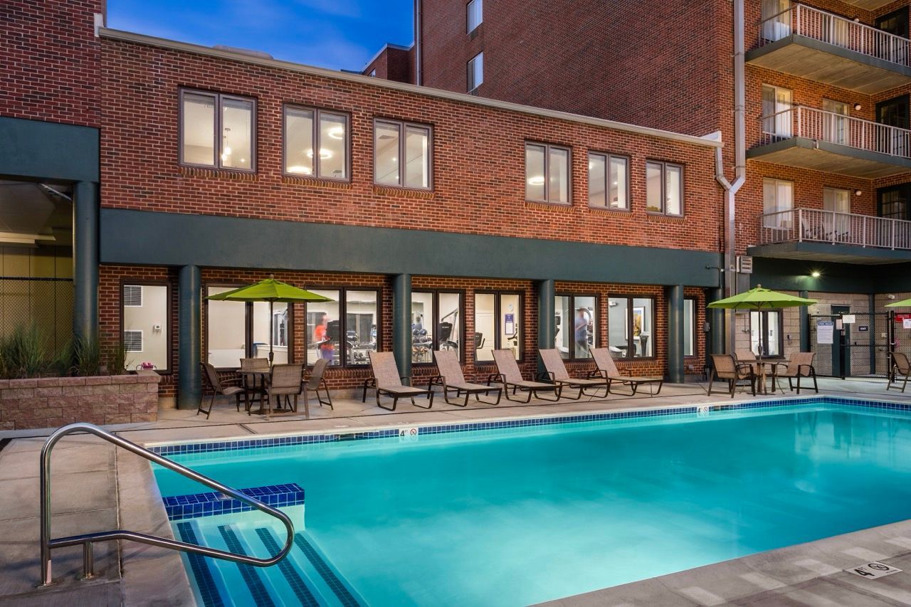 Outdoor pool area at a brick apartment community with lounge chairs and green umbrellas.