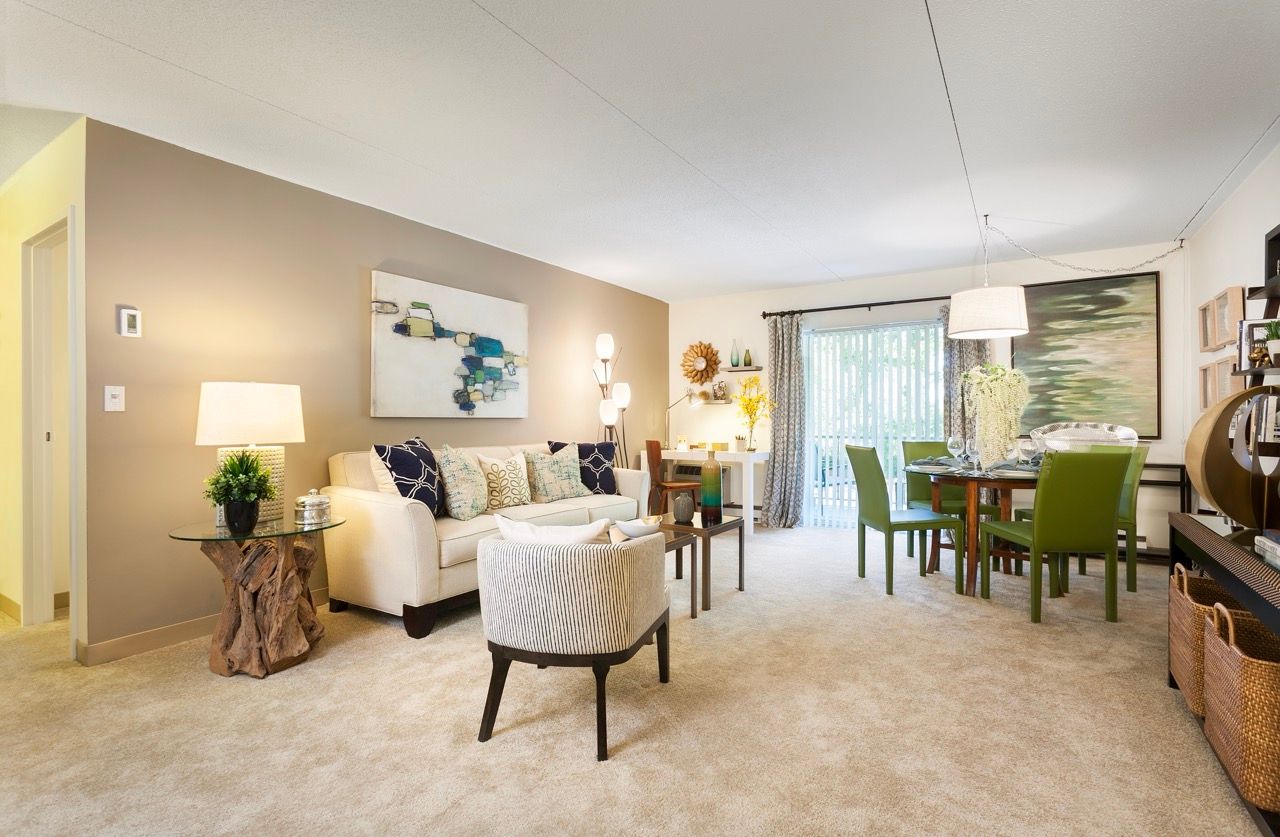 Open-plan living room in a modern apartment with beige carpet, sofa, chairs, and a dining table.