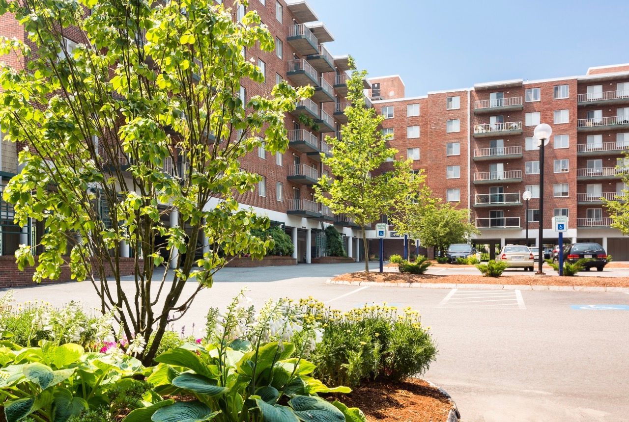 Exterior view of a brick apartment building with balconies, lush landscaping, and a parking lot.
