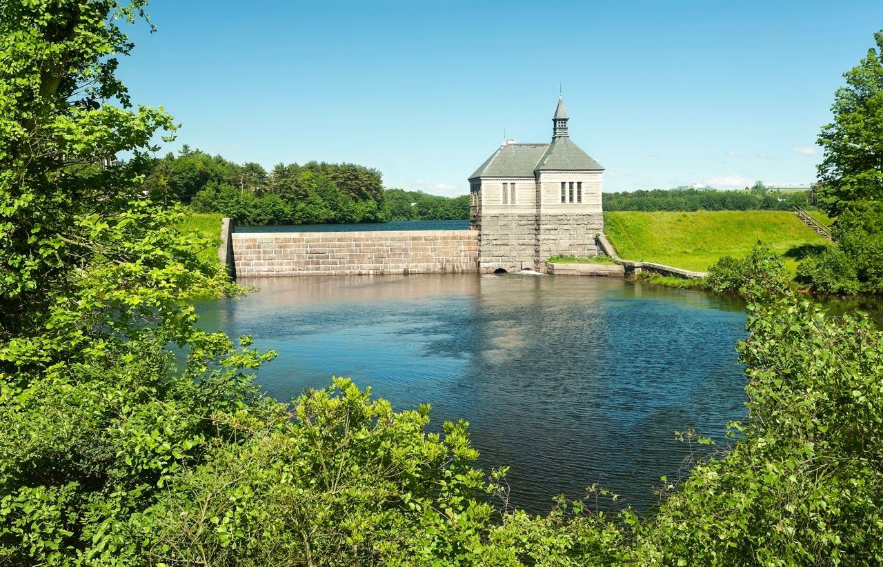 Historic stone dam with a small tower on the river, framed by green trees.