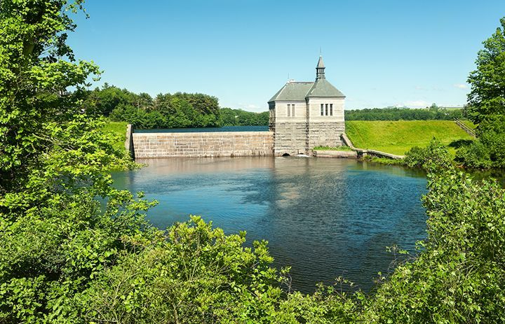 Stone water reservoir with a small building, calm blue water, and green trees under a clear sky