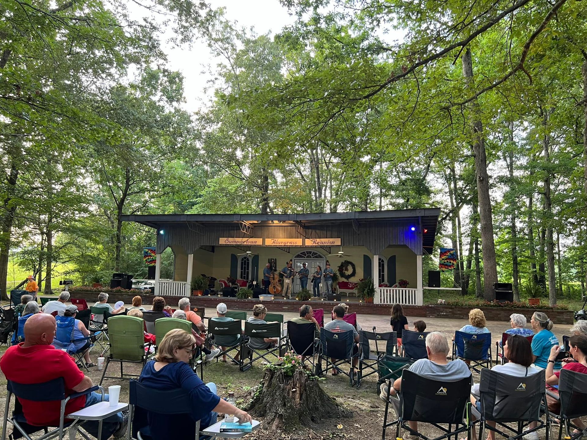 A group of people are sitting in chairs in front of a stage in the woods.