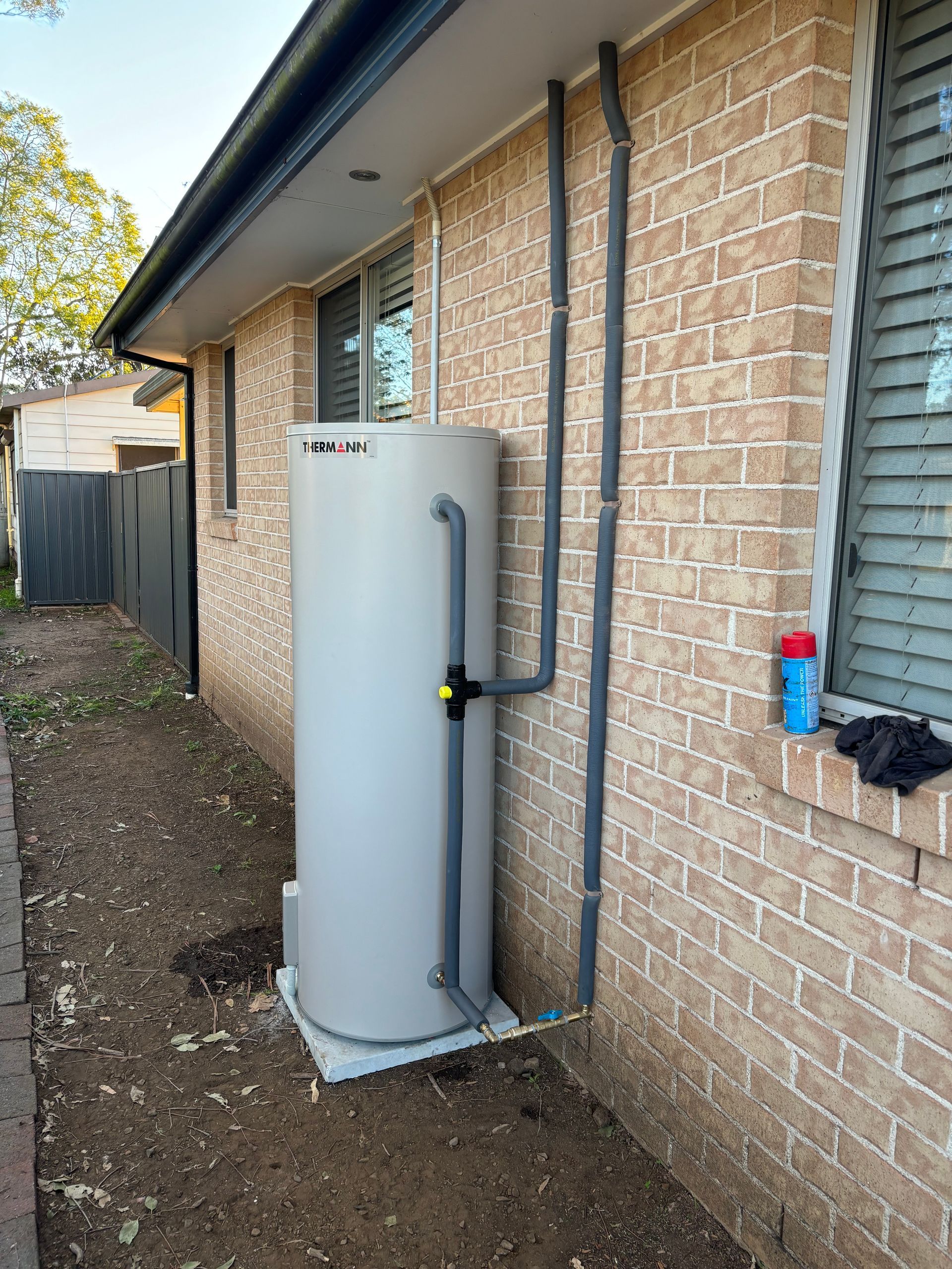 Water heater installed on a concrete slab against a brick wall, with exposed pipes and a nearby window.