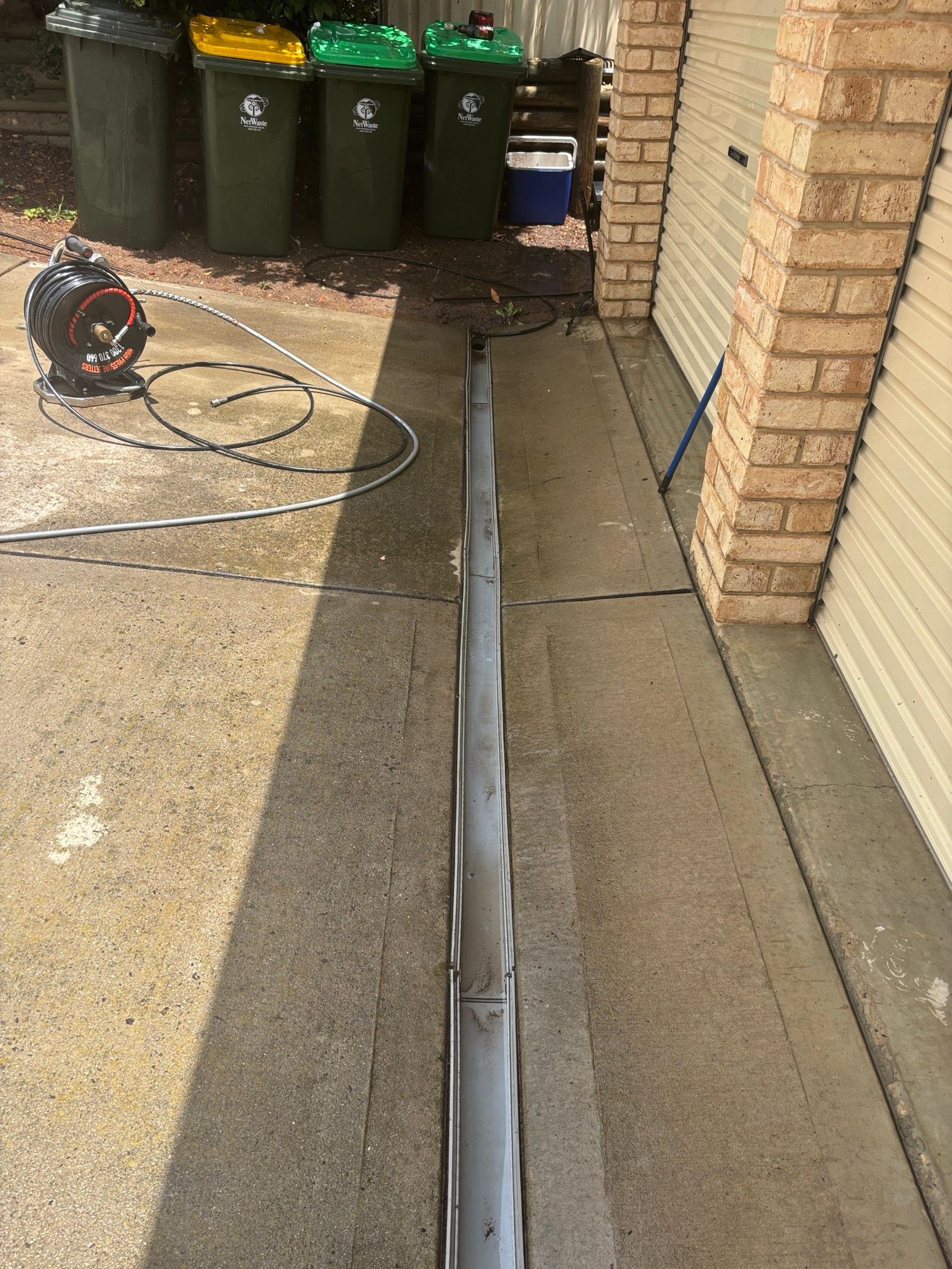 Concrete driveway with a drain being cleaned. A pressure washer and trash bins are in the background.