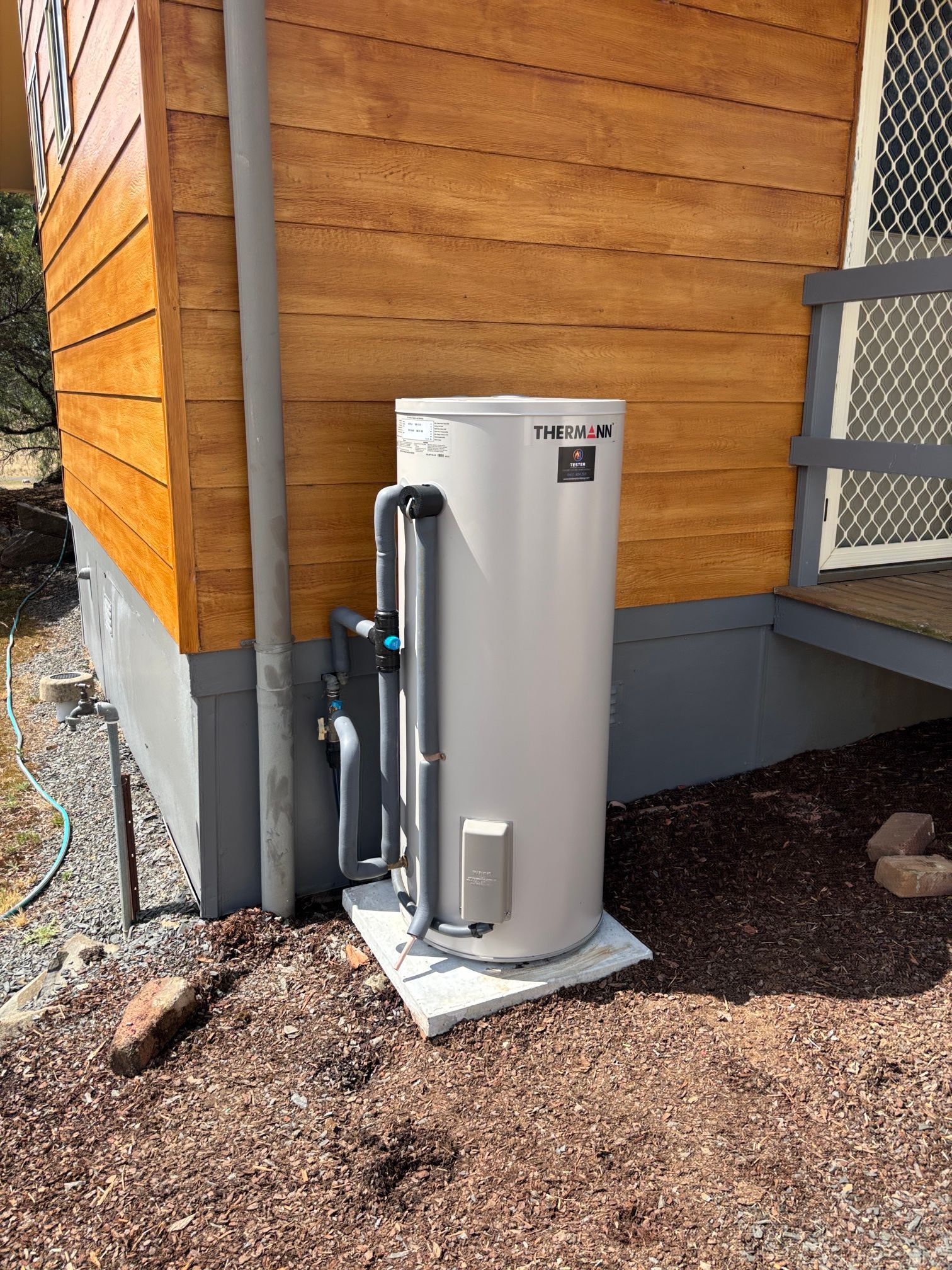 A cylindrical, light-colored hot water system outside a building with wooden siding, sitting on a concrete pad.