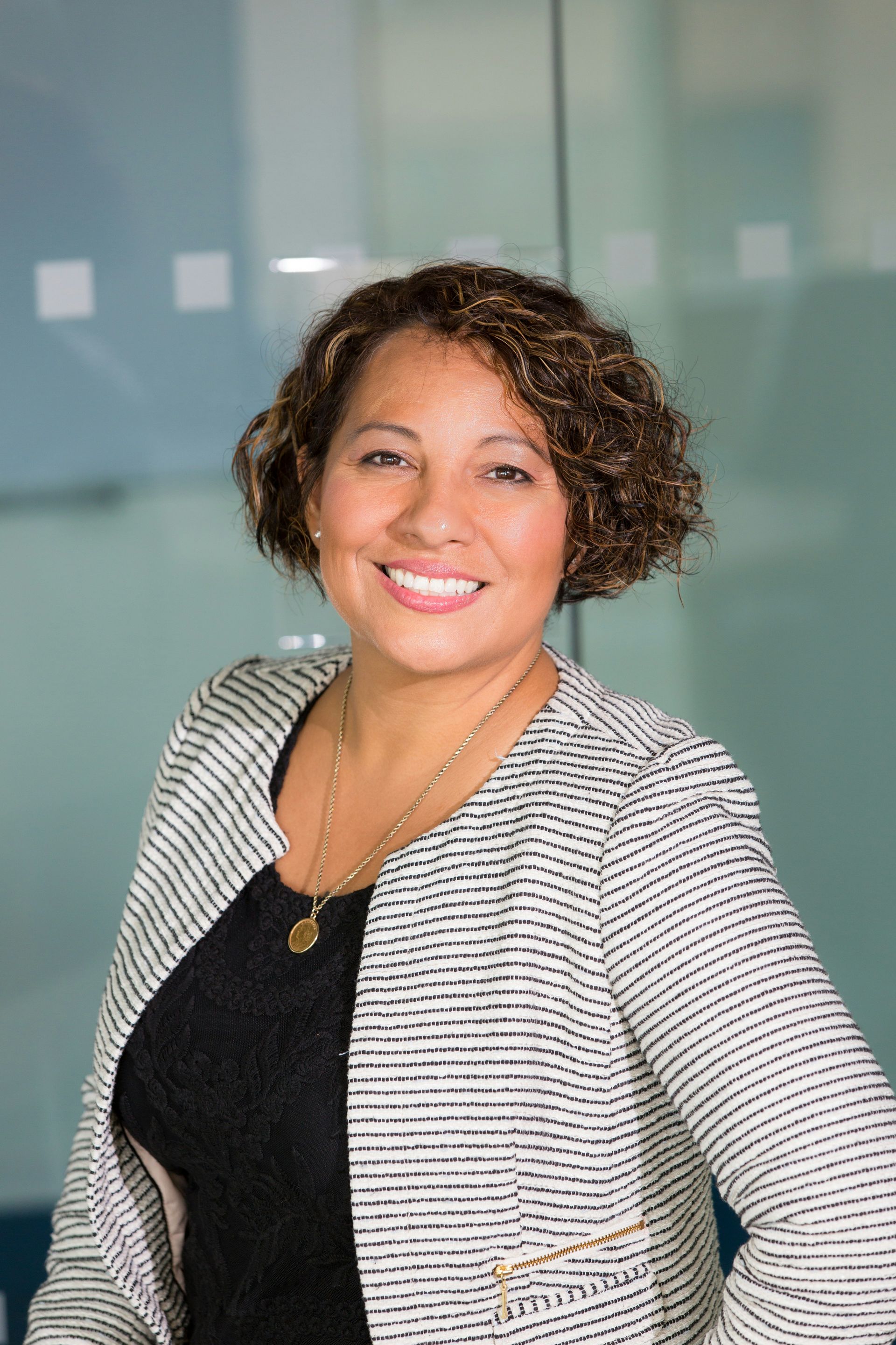 A person with short, curly hair smiling in a black top and patterned blazer against a glass-walled office background.