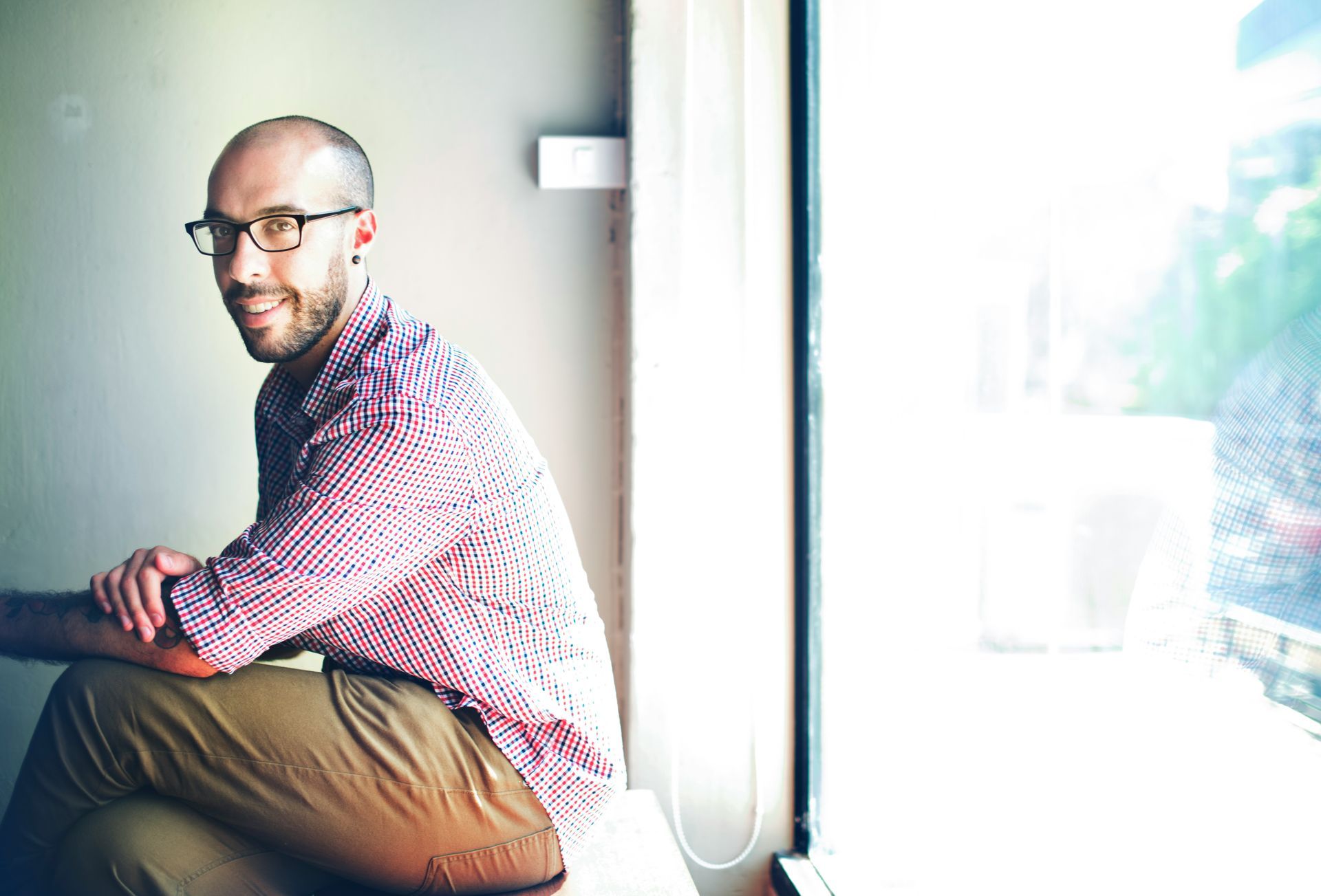 A person with a beard and glasses, wearing a checkered shirt and tan trousers, sits by a window and smiles at the camera.