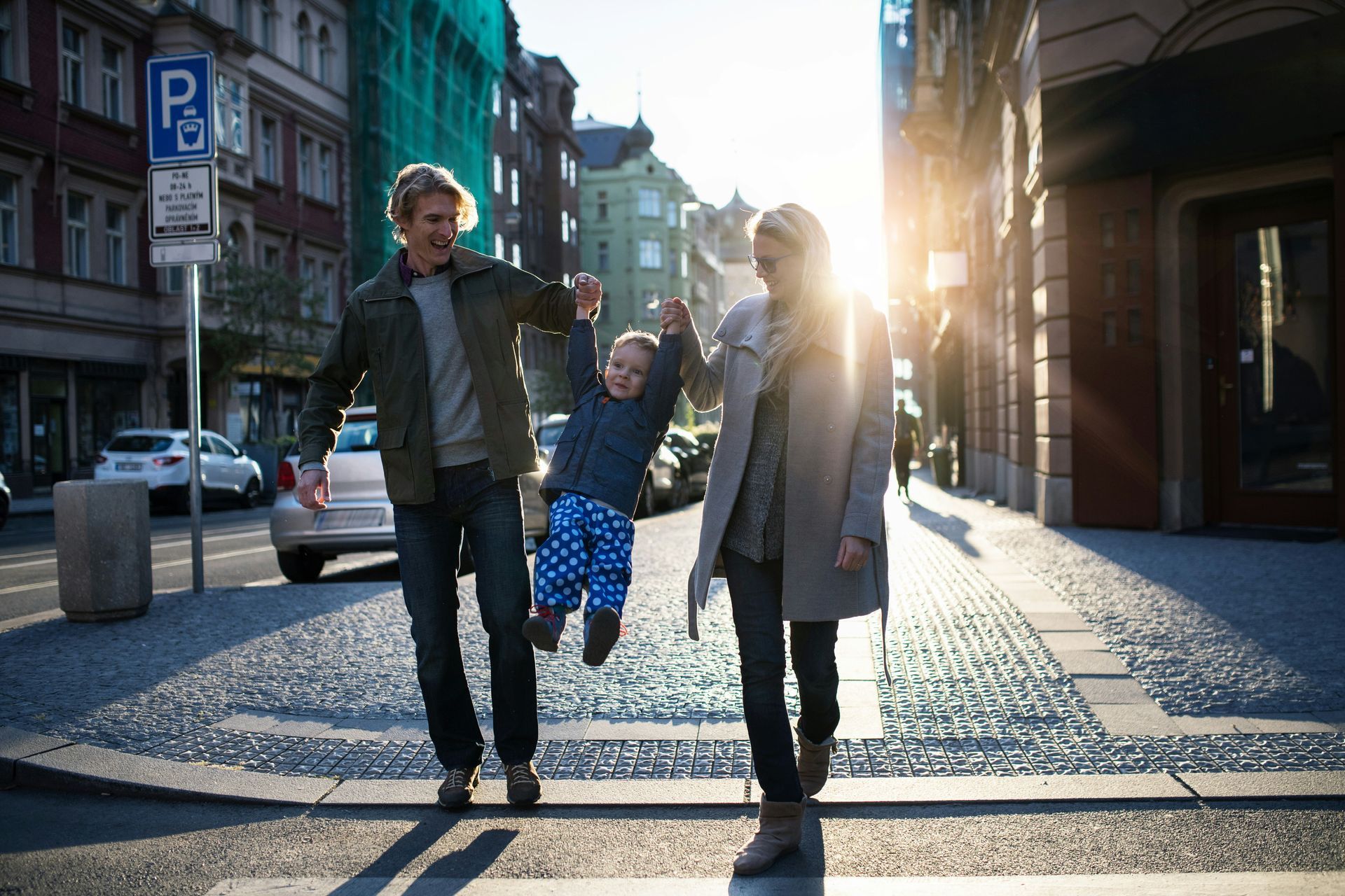 A family walking on a sunny city street, lifting a small child by the hands as they walk together across a crosswalk.
