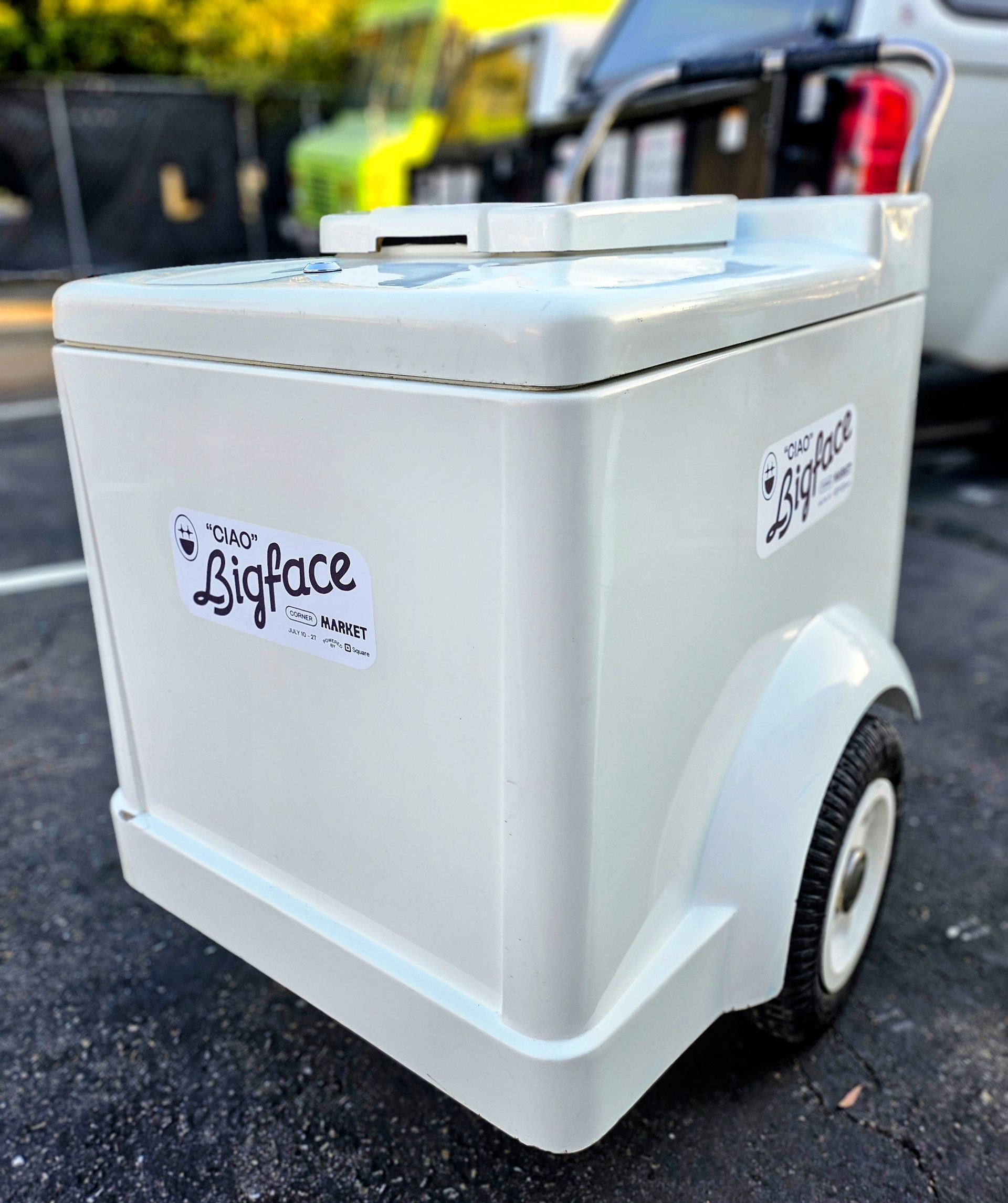 White portable cooler/cart with black wheels outdoors on pavement.