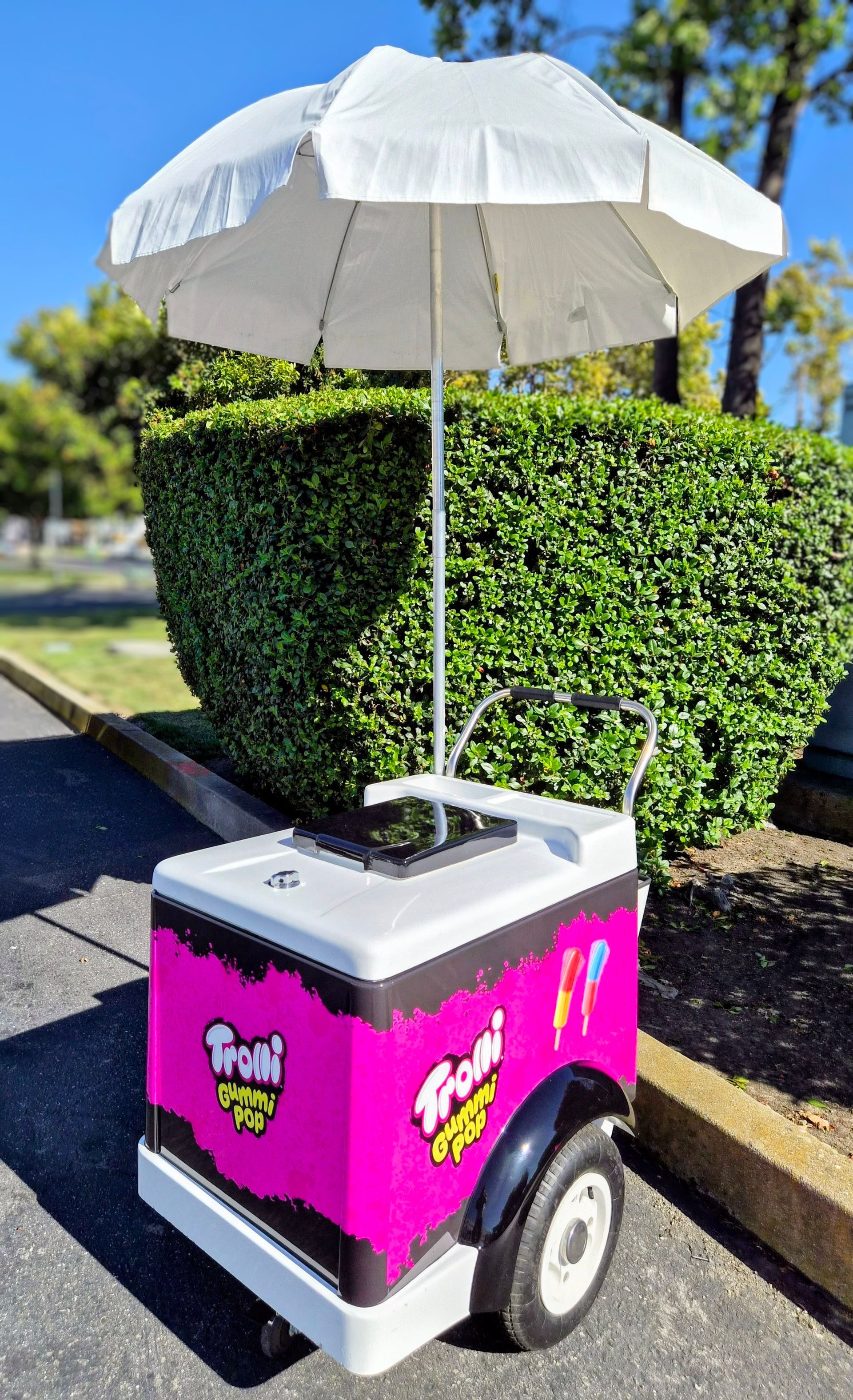 White umbrella on a pink and black cart parked beside a hedge on a sunny street.