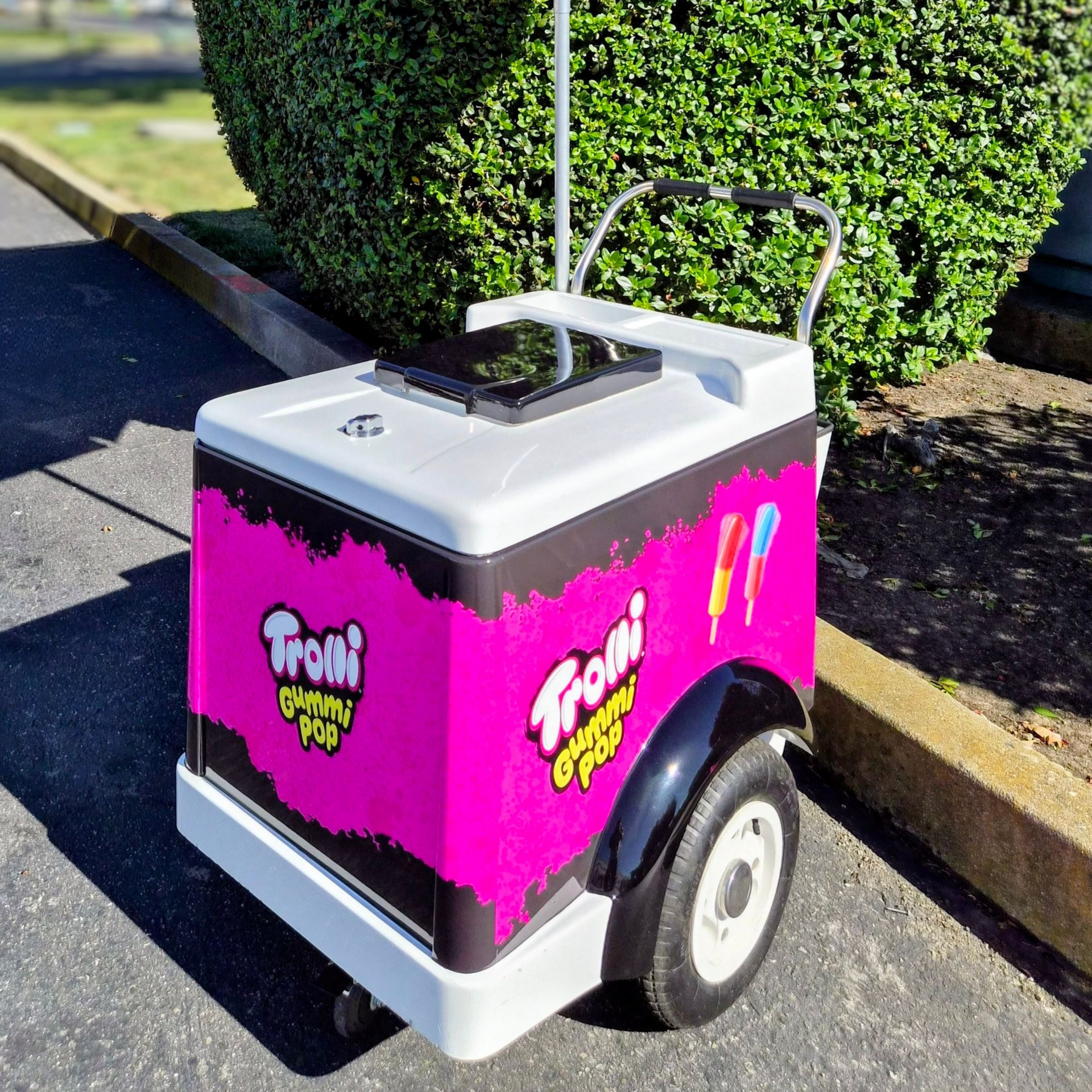 Pink and white cooler cart with handle and wheels parked on pavement beside a hedge