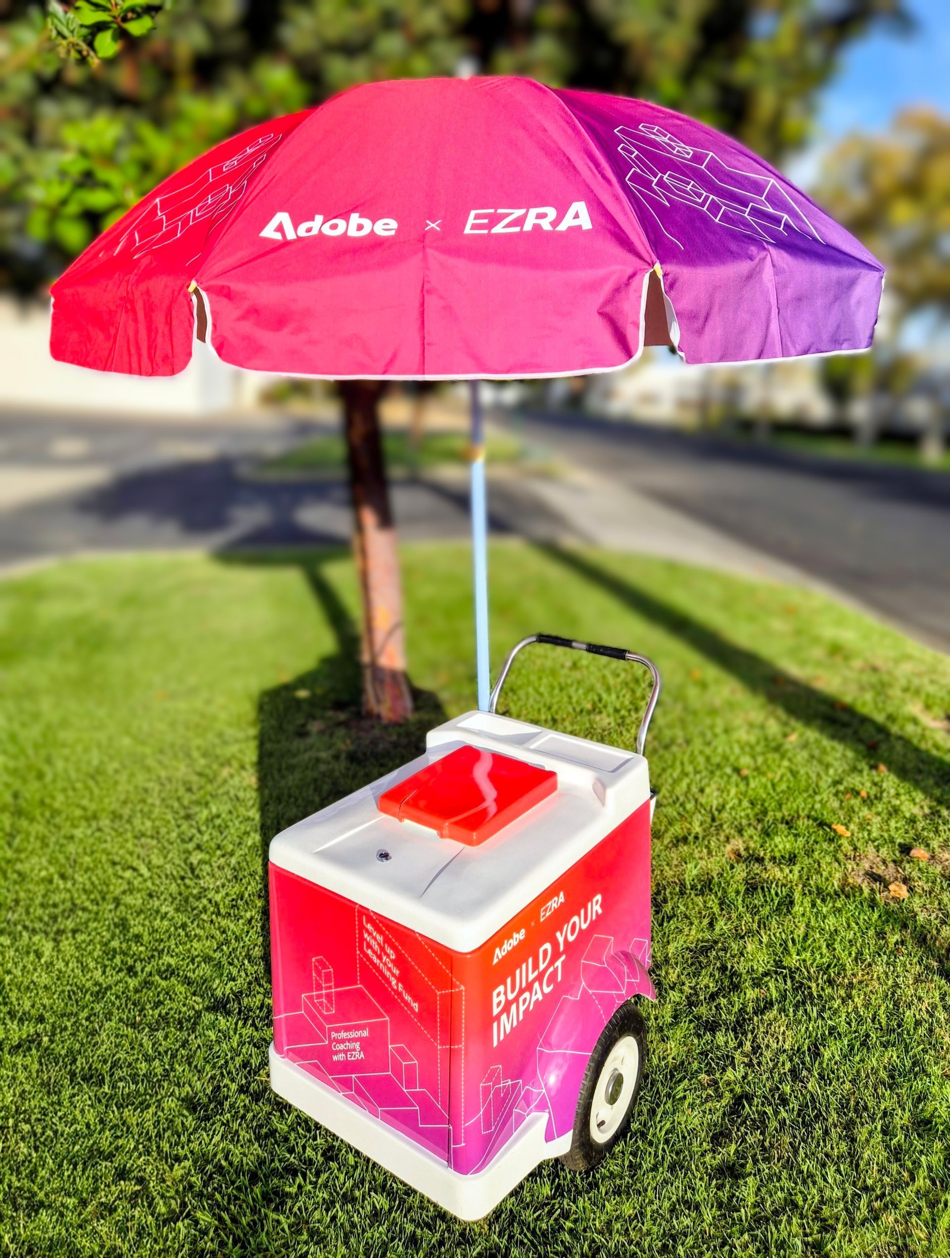 Pink and white EZRA umbrella shade over a small pink cooler cart on grass outdoors.