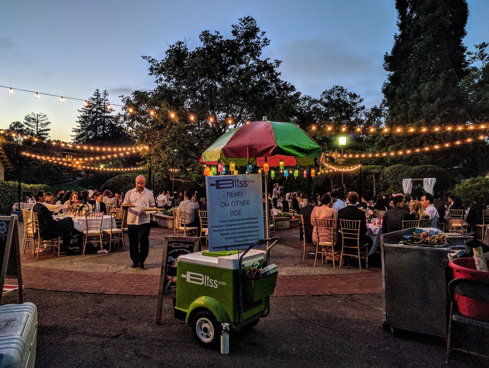 A green ice cream cart is parked in front of a crowd of people at a party.