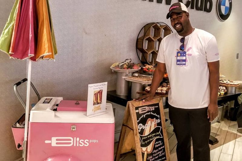 Man posing beside a pink Bisss booth with drinks and a menu sign at an indoor event