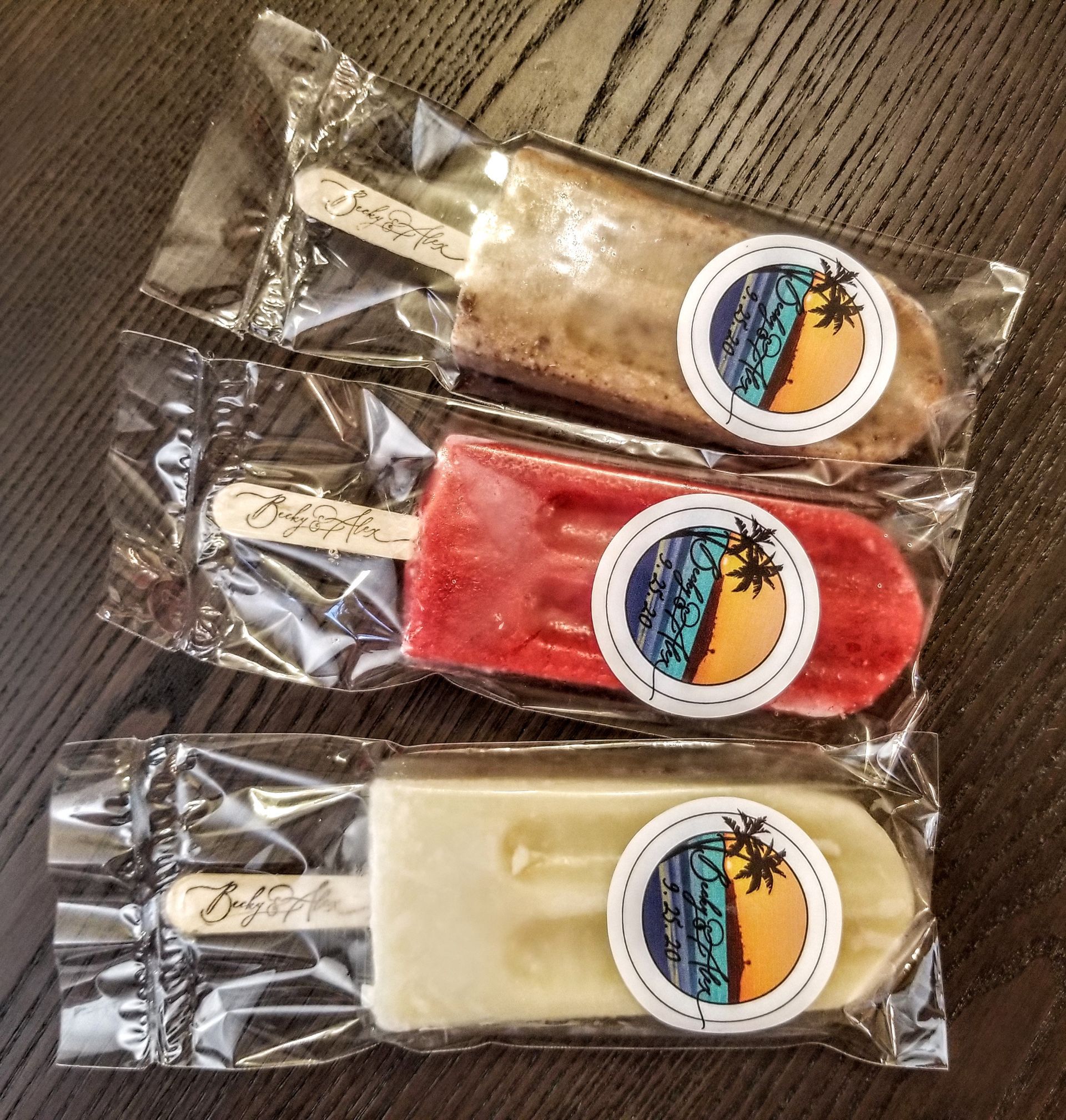Three popsicles are wrapped in plastic and sitting on a wooden table.