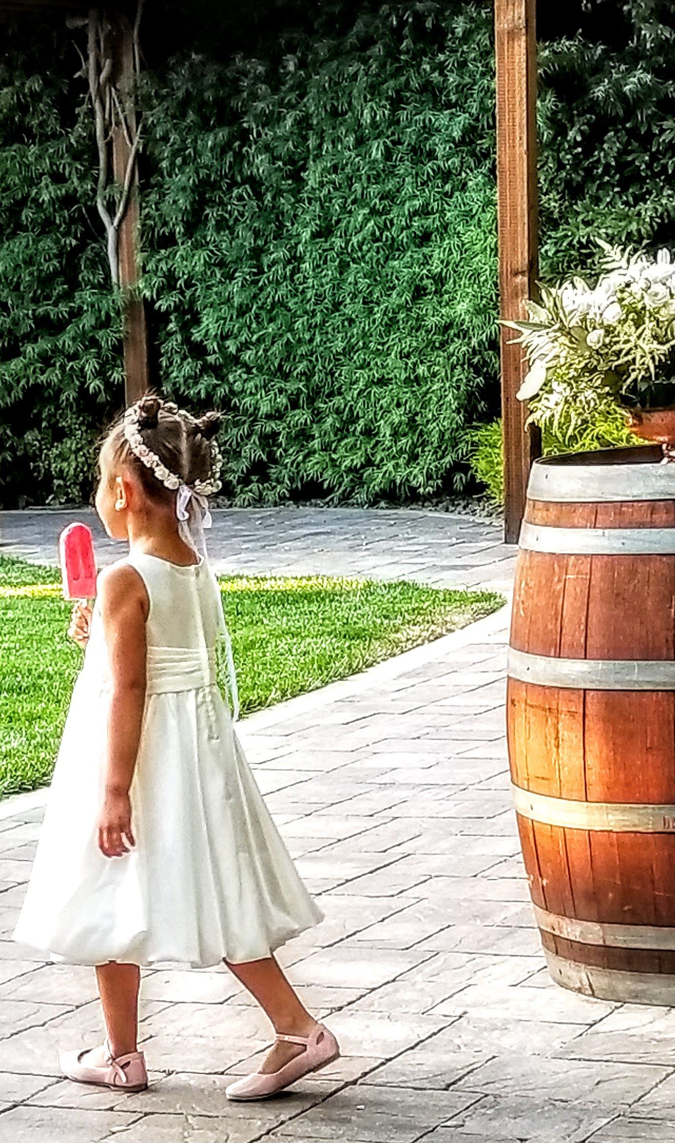 A little girl in a white dress is walking down a sidewalk next to a wooden barrel.