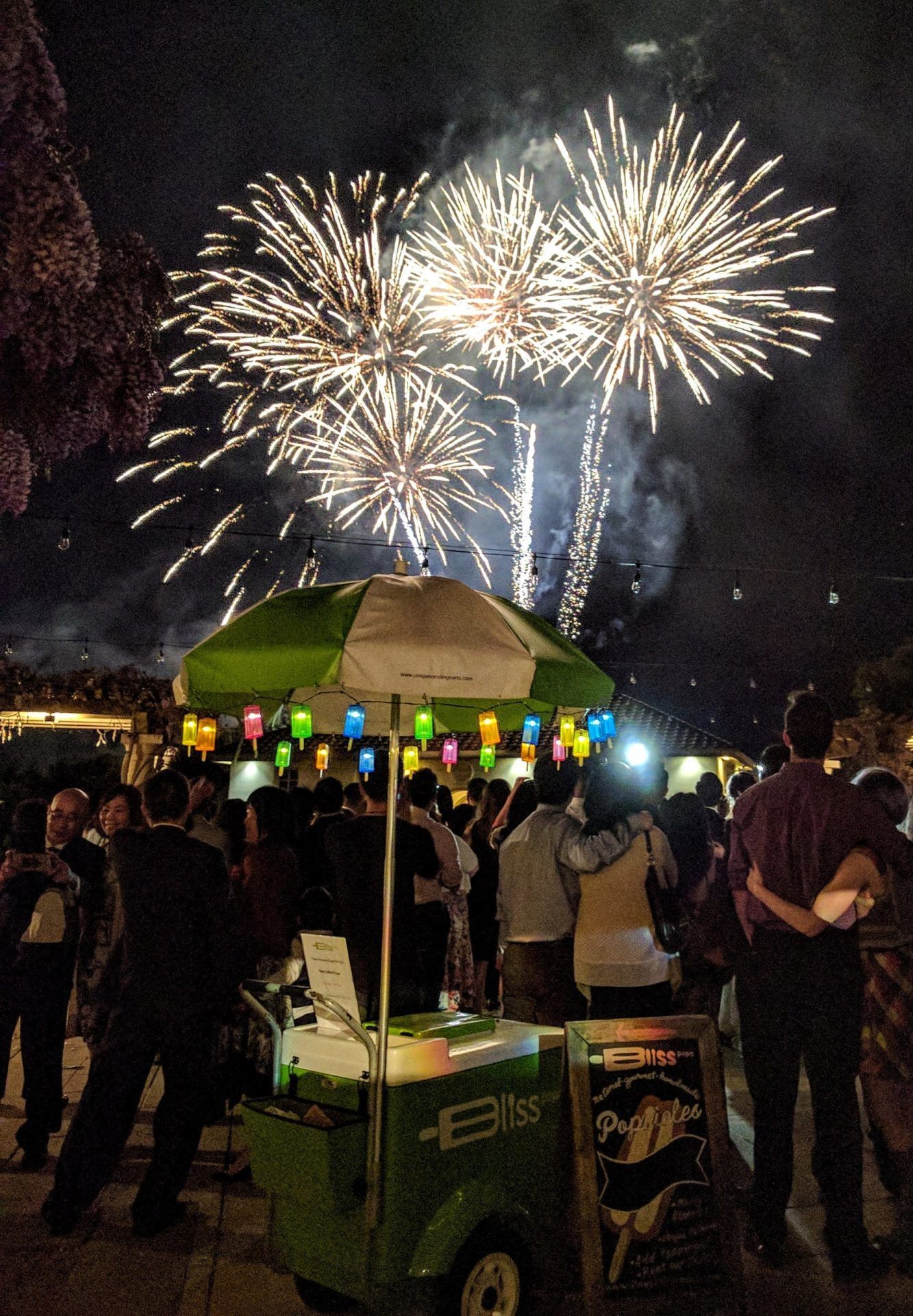 A green and white ice cream cart is surrounded by people watching fireworks