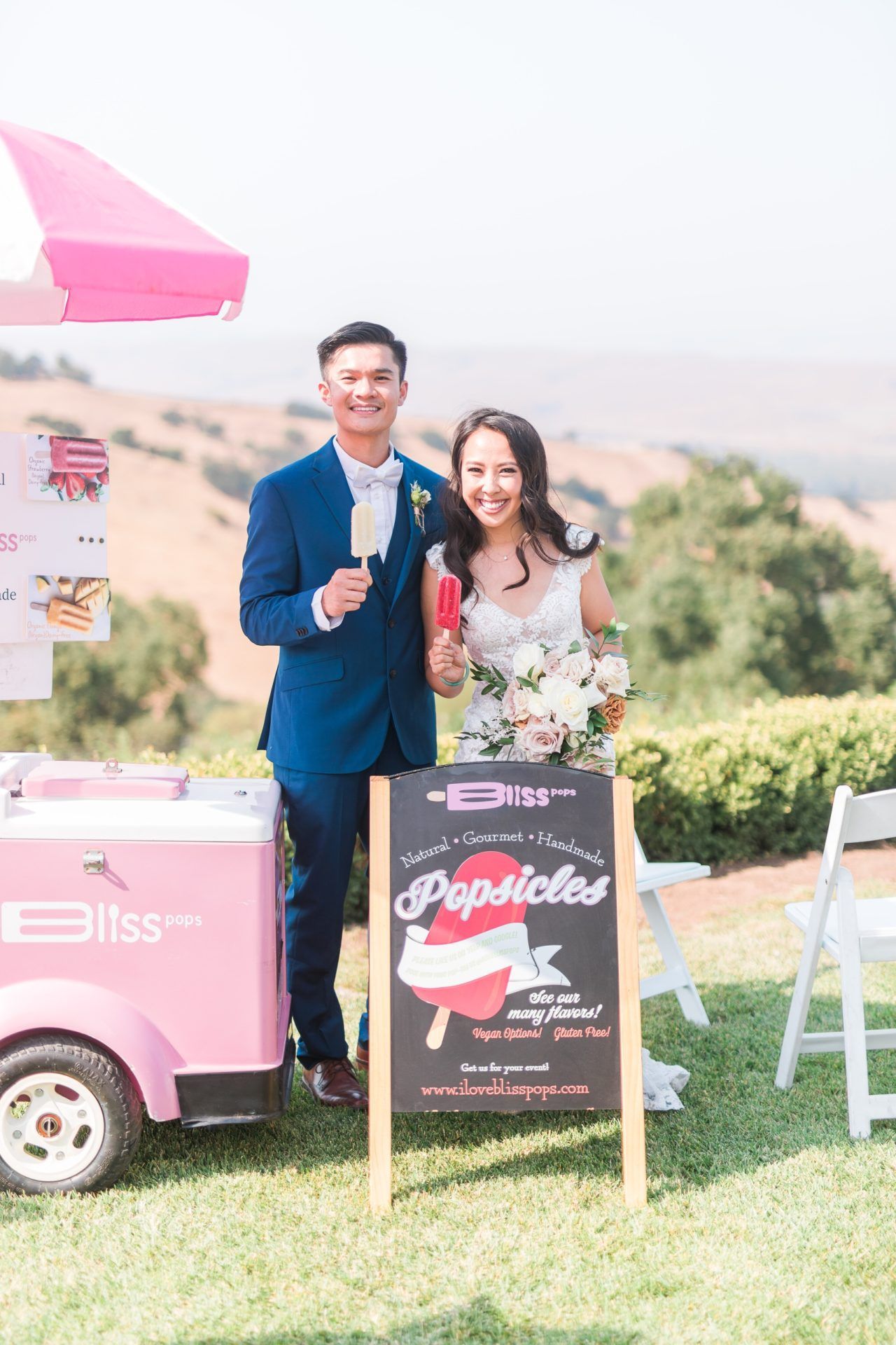 A bride and groom are standing in front of an ice cream cart.