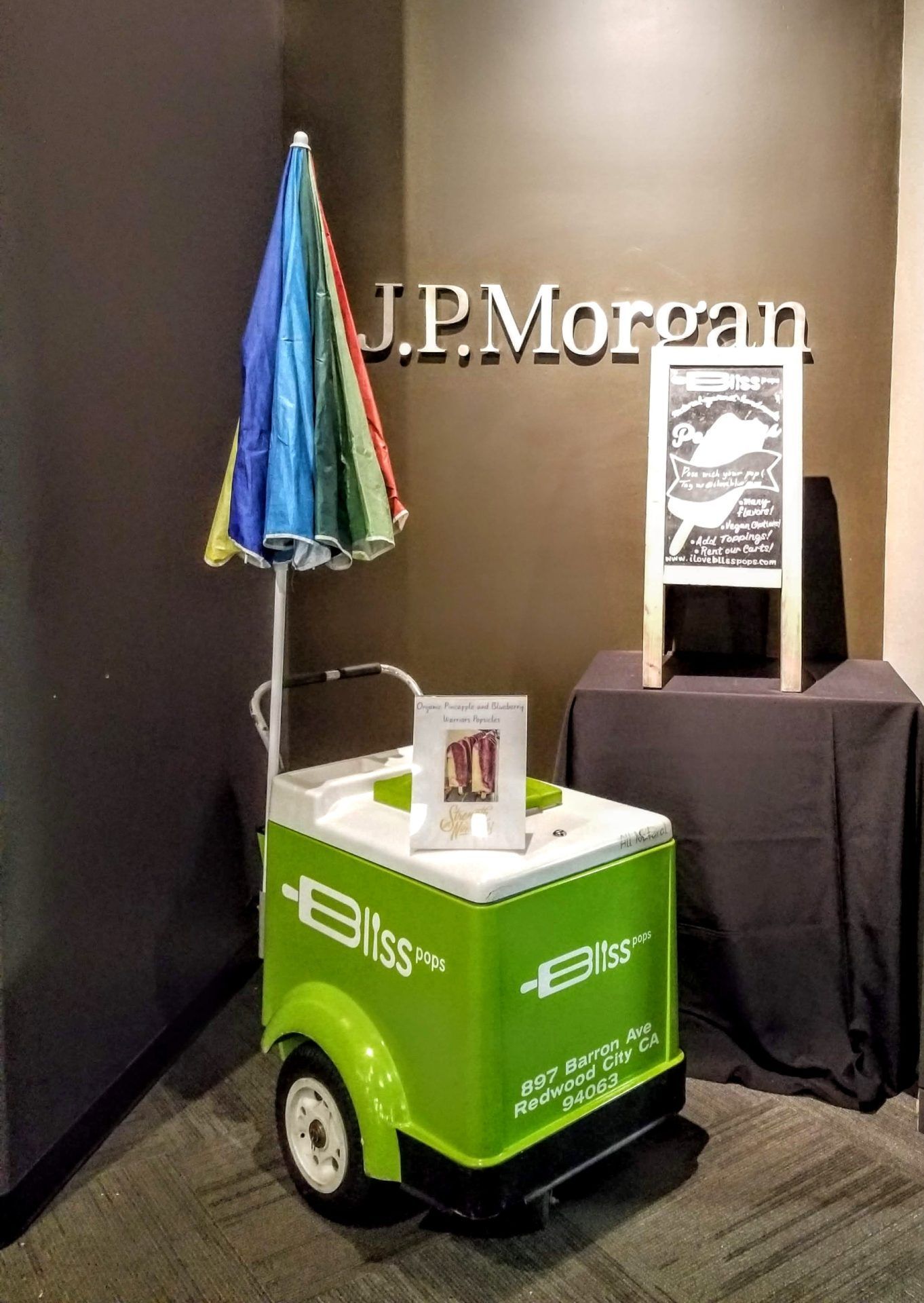 A green and white ice cream cart with an umbrella in front of a sign that says j.p. morgan.