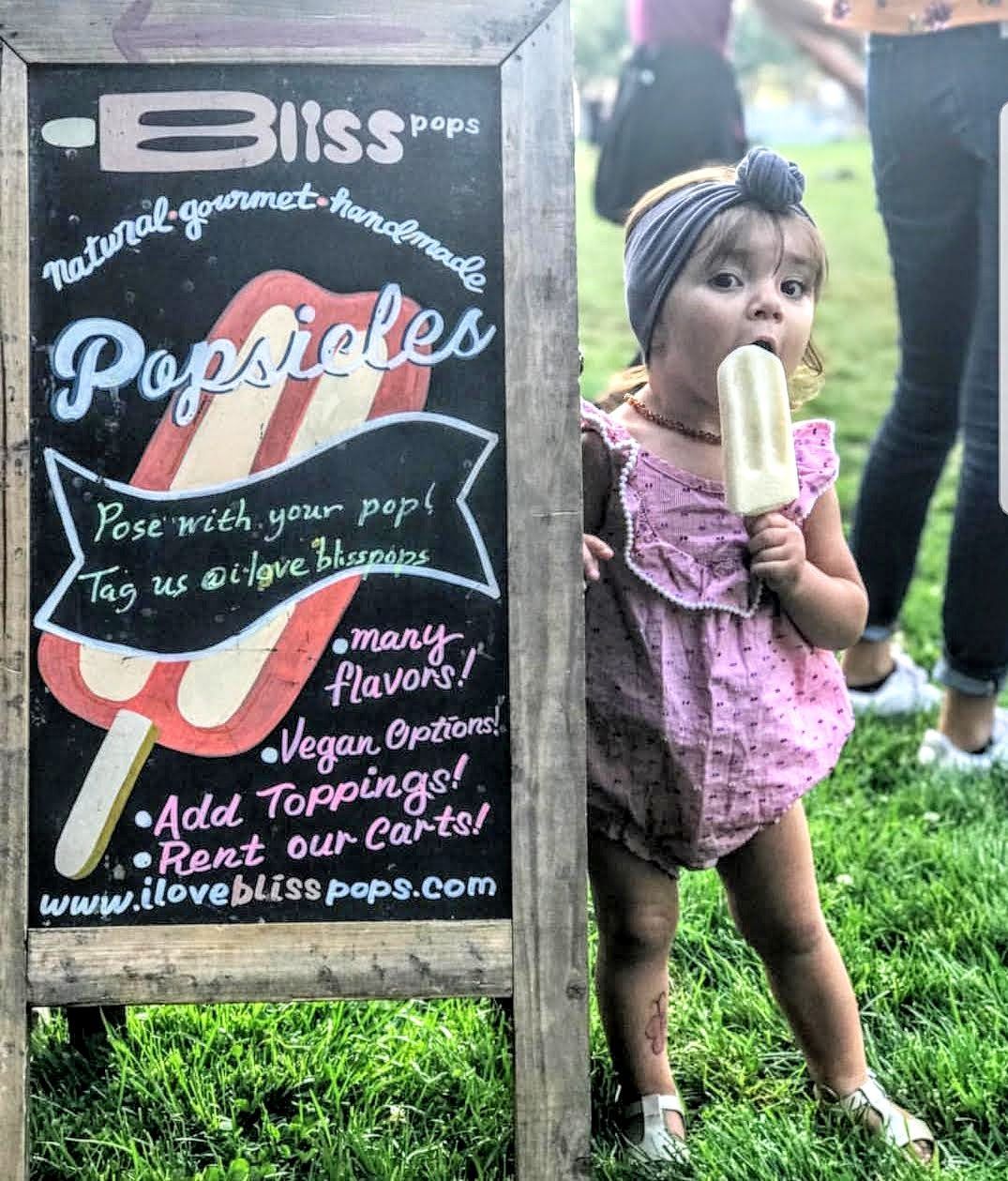 A little girl is eating a popsicle in front of a sign that says popsicles.