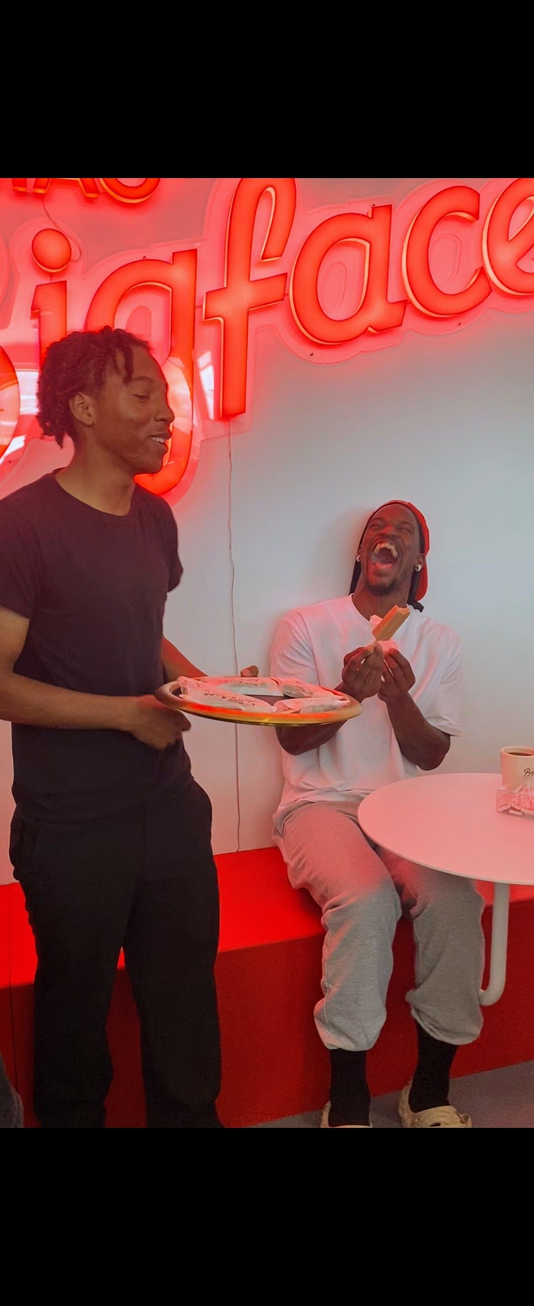 Two people in a red-lit neon lounge, one standing with a tray and one seated by a white table.