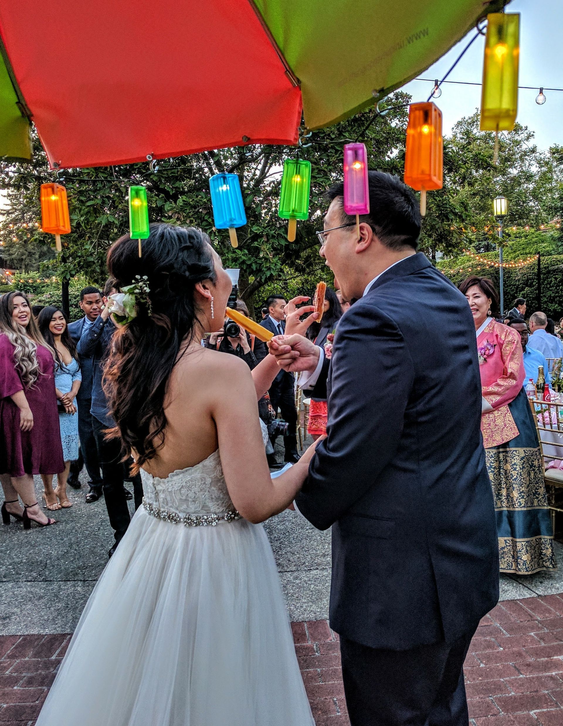 A bride and groom are cutting their wedding cake under a colorful umbrella.