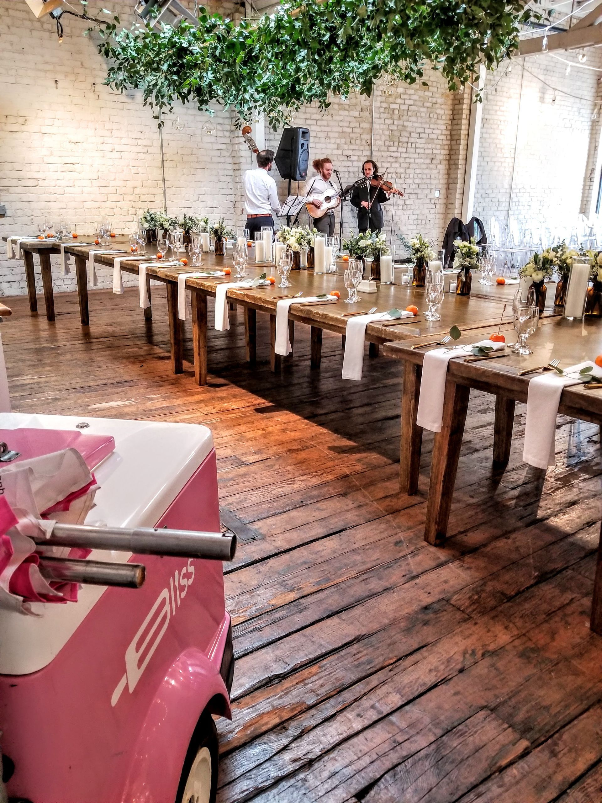 A pink ice cream cart is parked in front of a long table set for a wedding reception.