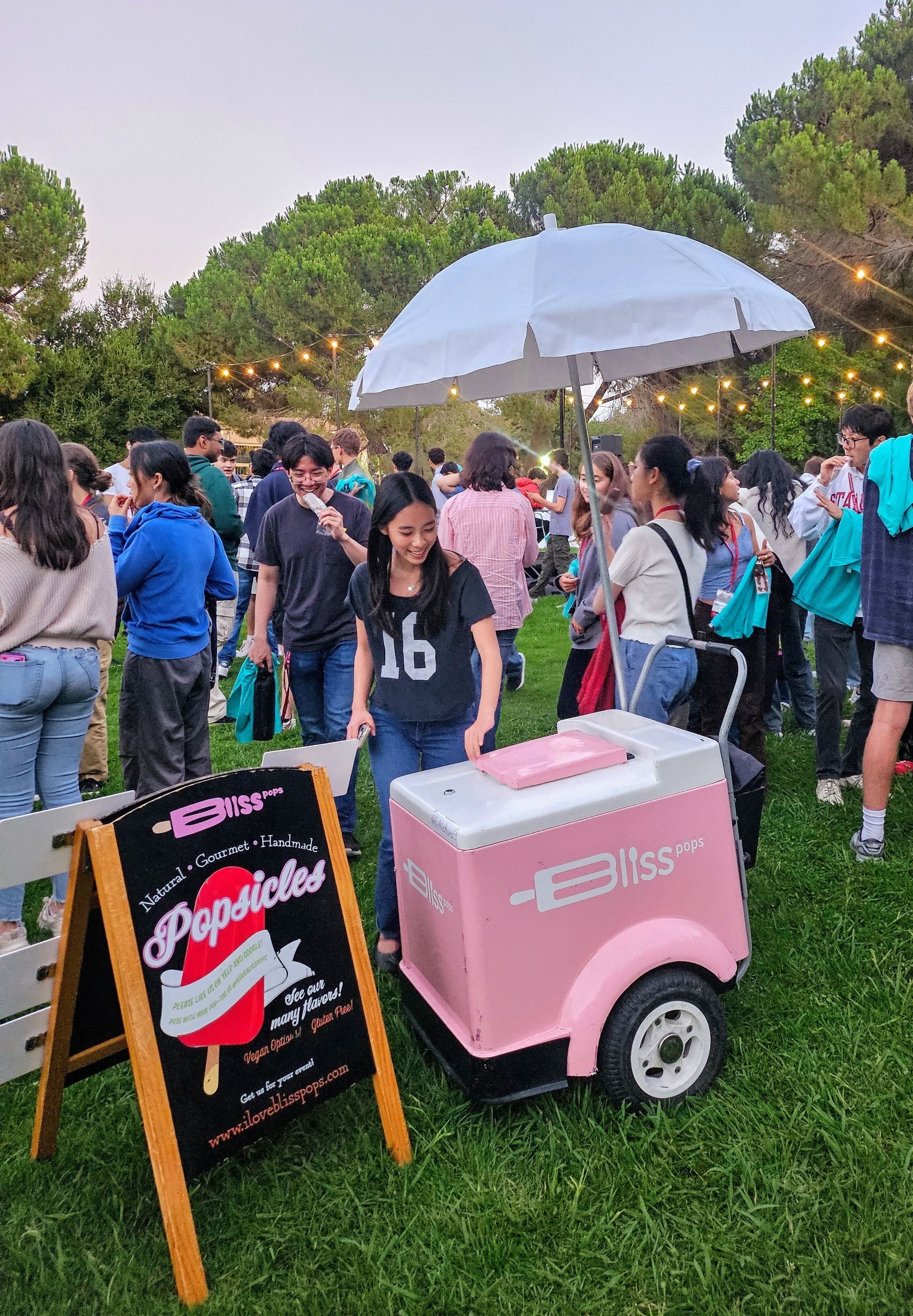 Crowd at outdoor event with pink ice cream cart, chalkboard sign, and white umbrella on a grassy lawn