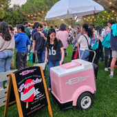 Crowd at outdoor event with pink ice cream cart and white umbrella on a grassy field
