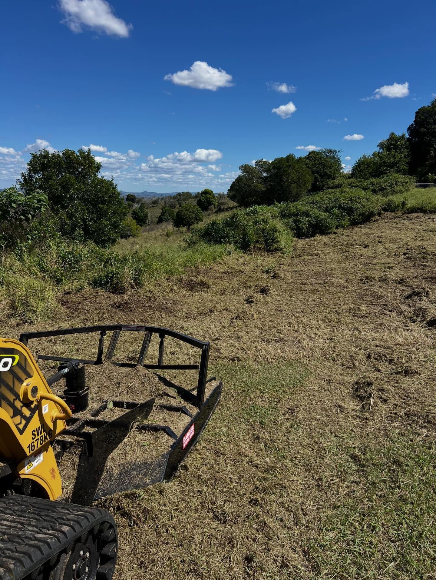 A Yellow Tracked Machine Clearing Brush on a Grassy Hillside Under a Blue Sky — Dudgeon’s Earthworks in Murgon, QLD