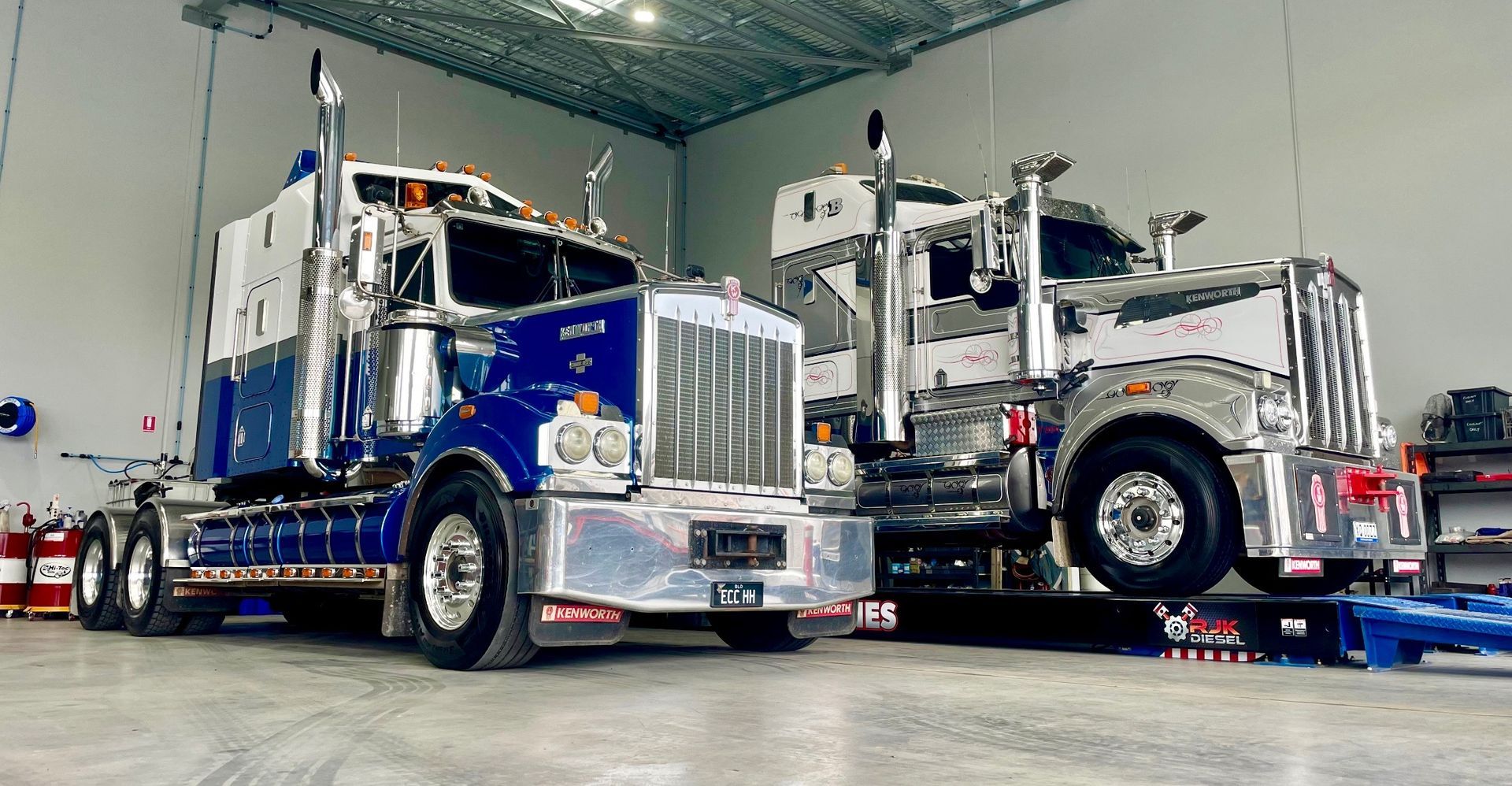 Two shiny semi-trucks, one blue and white, the other silver and white, parked in a well-lit garage — ECC Heavy Haulage in Ormeau, QLD