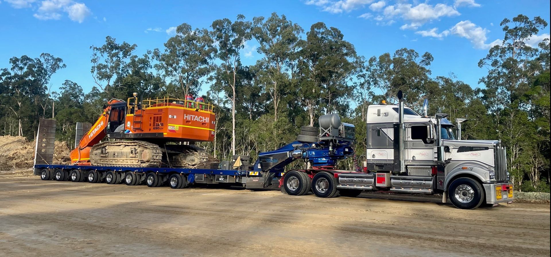 An excavator on a trailer being pulled by a semi-truck in a wooded area — ECC Heavy Haulage in Ormeau, QLD