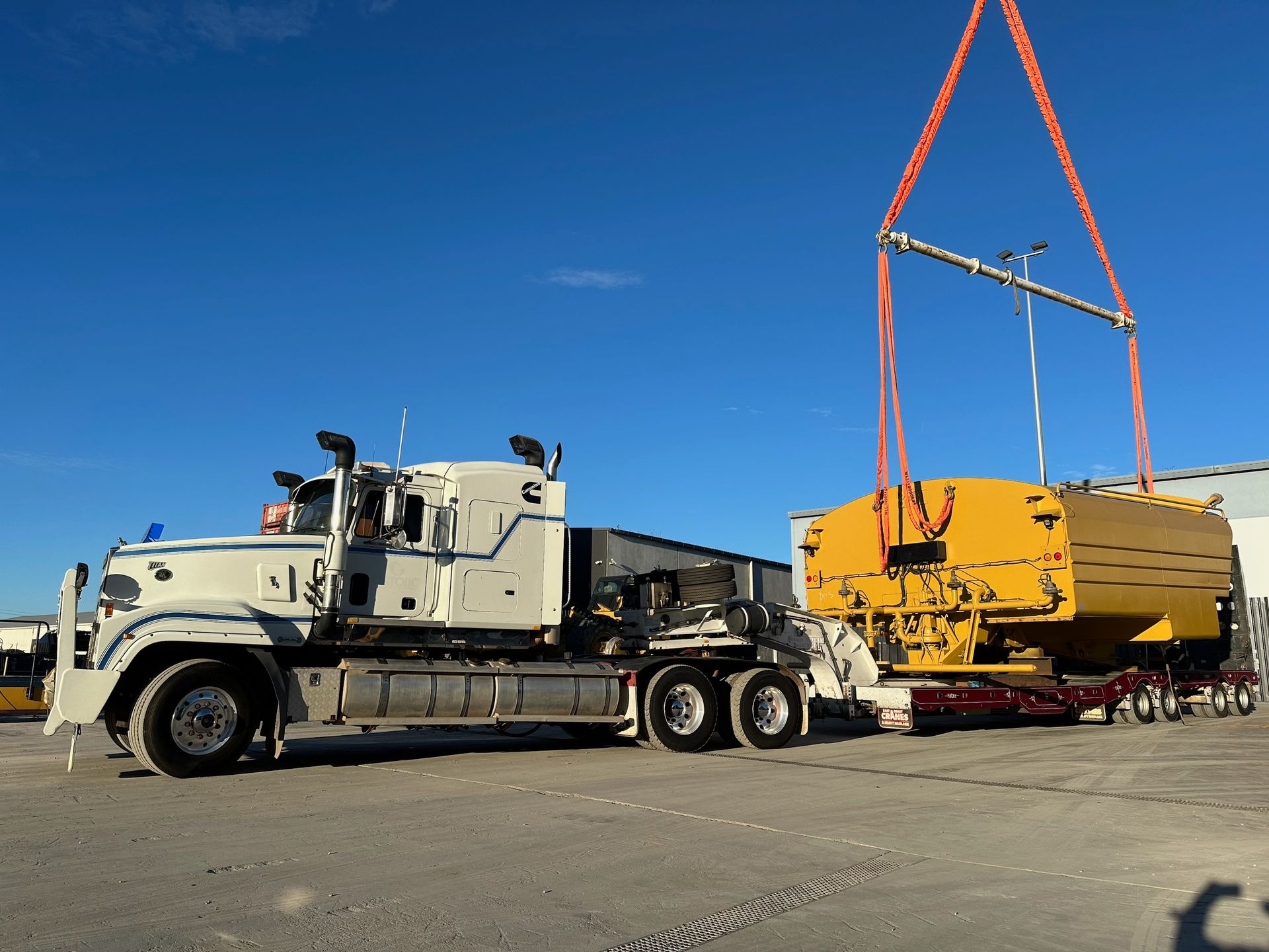White truck hauling yellow industrial equipment under a crane — ECC Heavy Haulage in Ormeau, QLD