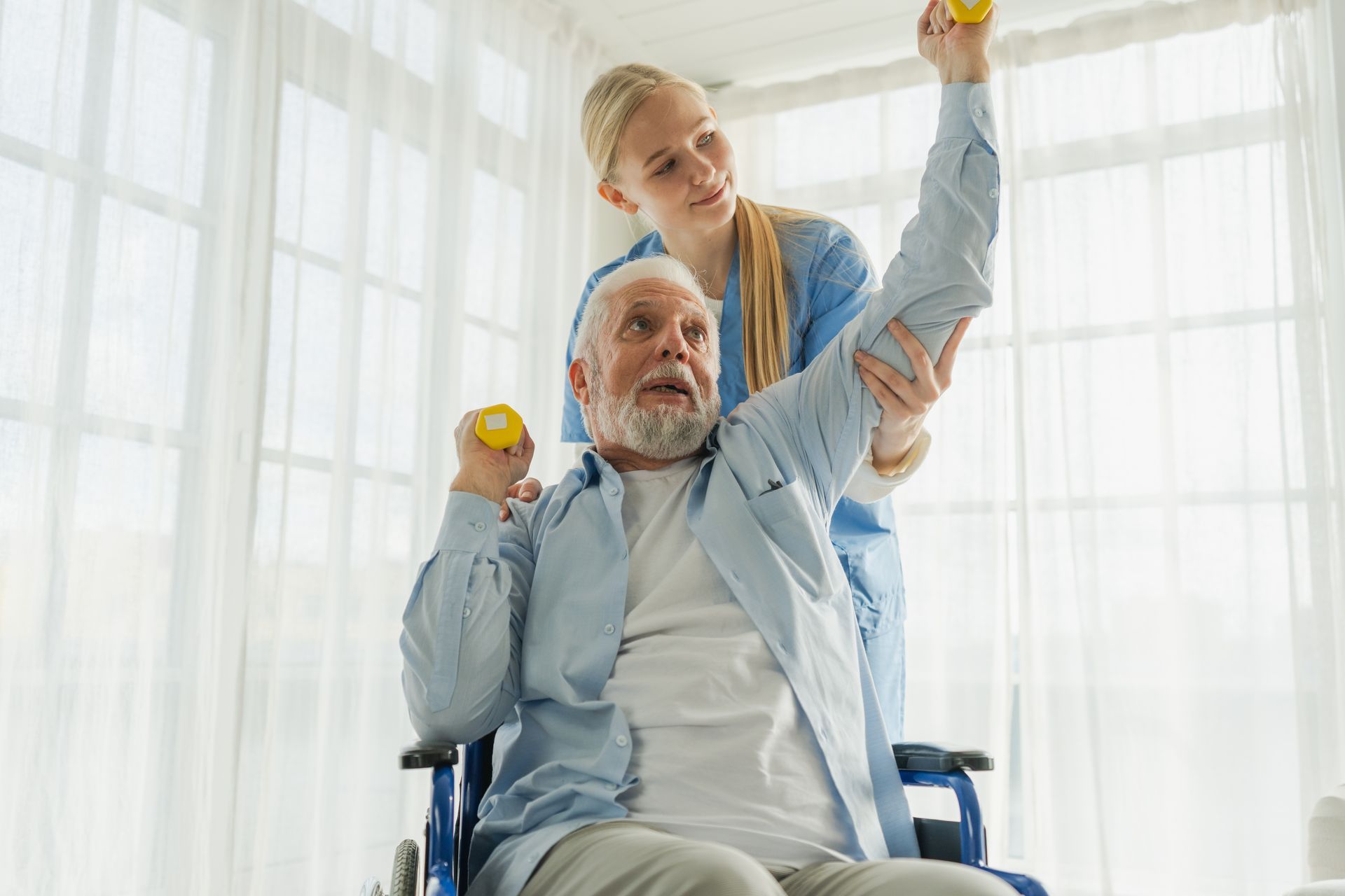 A caregiver in blue scrubs assists a person in a wheelchair with arm exercises using small yellow weights in a bright room. A caregiver in blue scrubs assists a person in a wheelchair with arm exercises using small yellow weights in a bright room.