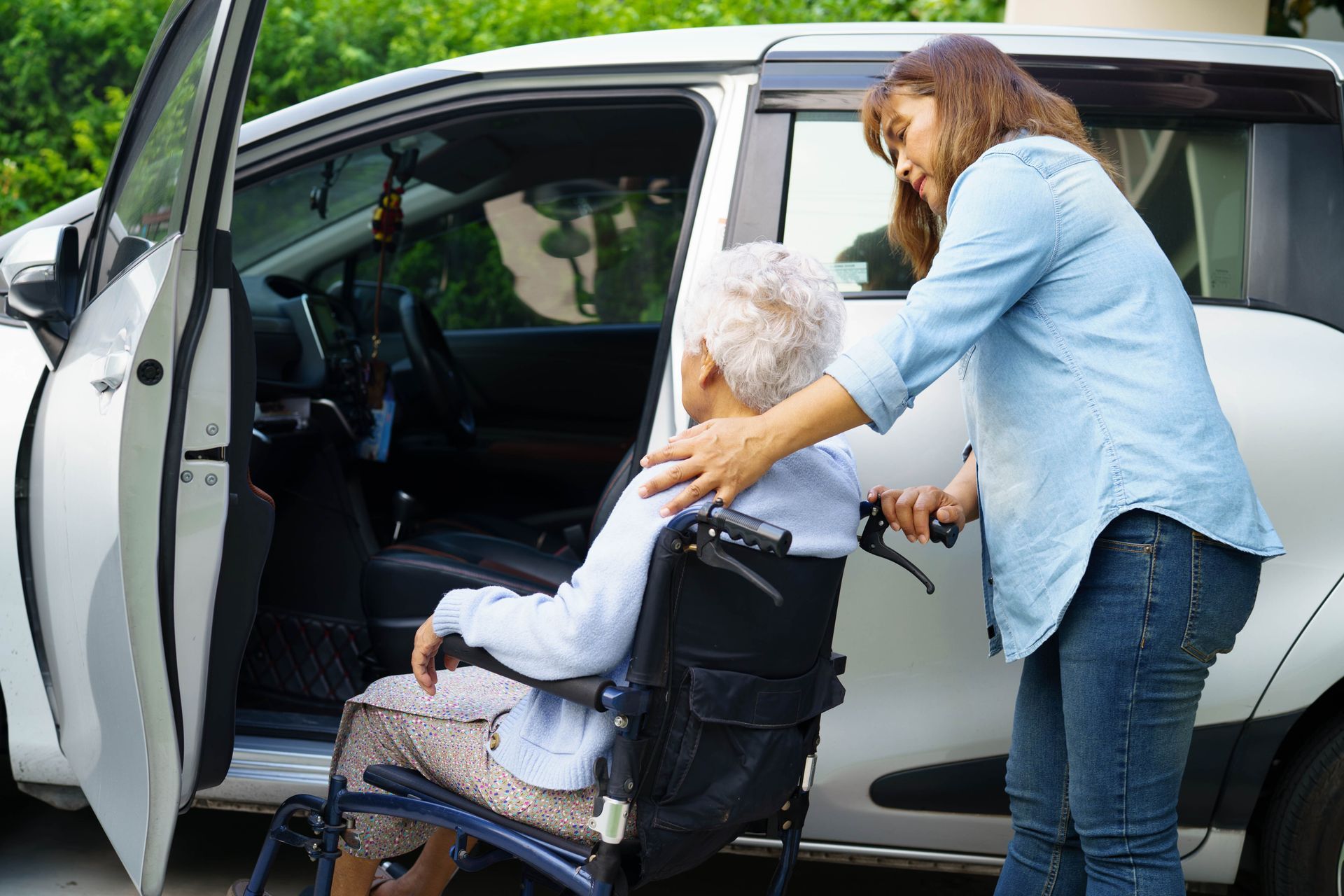 A caregiver helps a person in a wheelchair transfer into the front seat of a silver car. A caregiver helps a person in a wheelchair transfer into the front seat of a silver car.