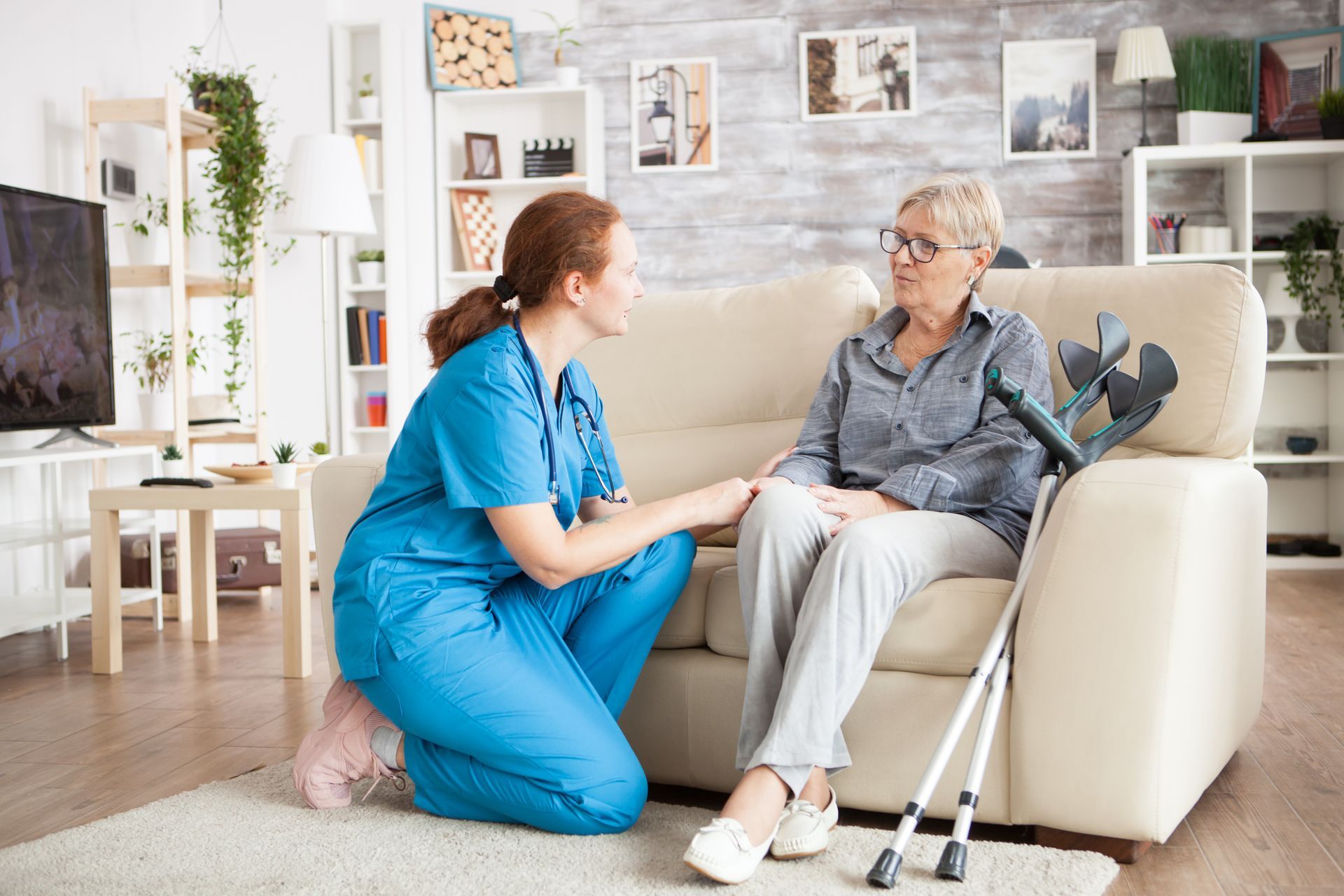 A medical professional in blue scrubs kneels by a couch, holding the hand of a patient with crutches in a living room. A medical professional in blue scrubs kneels by a couch, holding the hand of a patient with crutches in a living room.