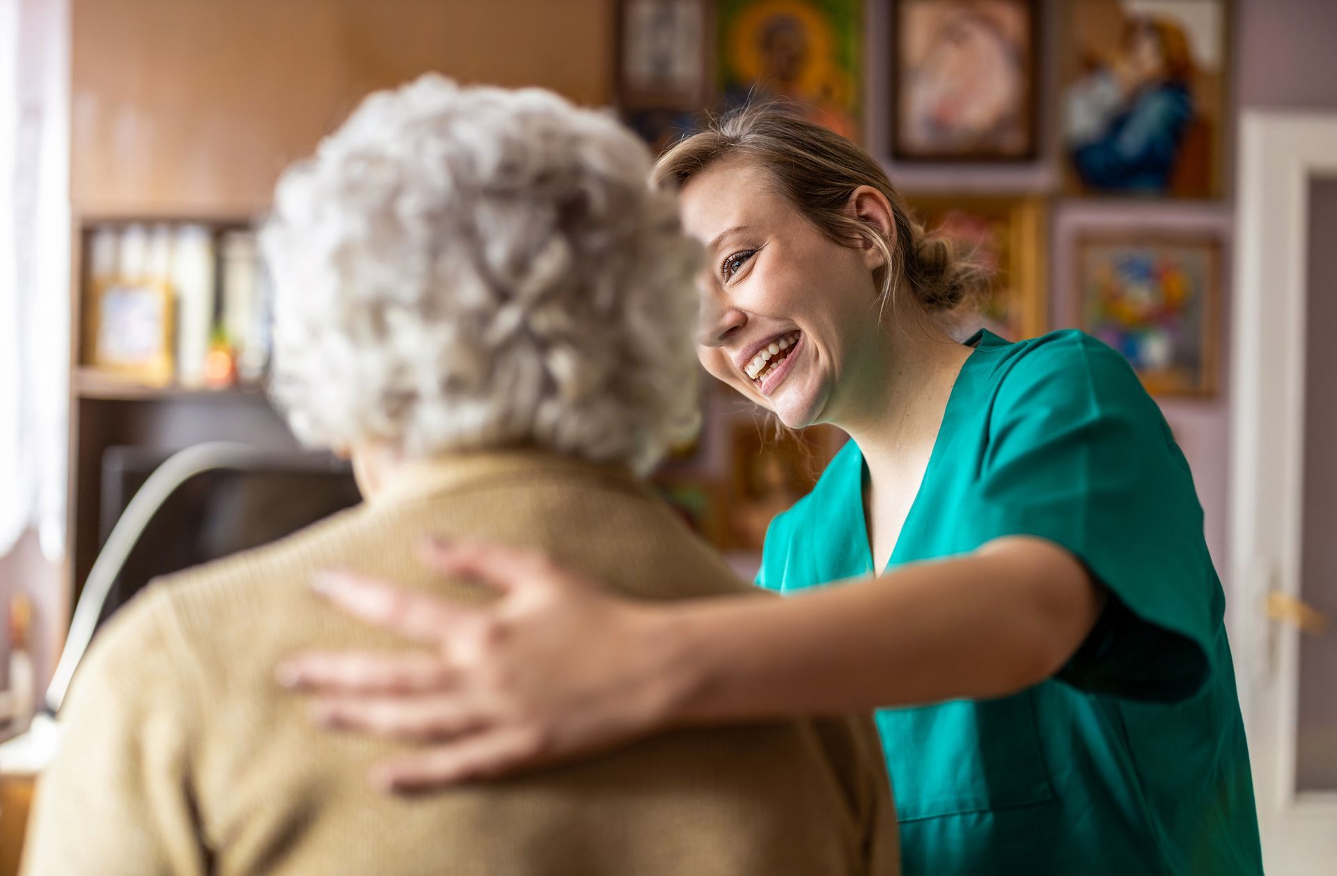 A caregiver in a green uniform smiling warmly while placing a supportive hand on the shoulder of a person with gray hair. A caregiver in a green uniform smiling warmly while placing a supportive hand on the shoulder of a person with gray hair.