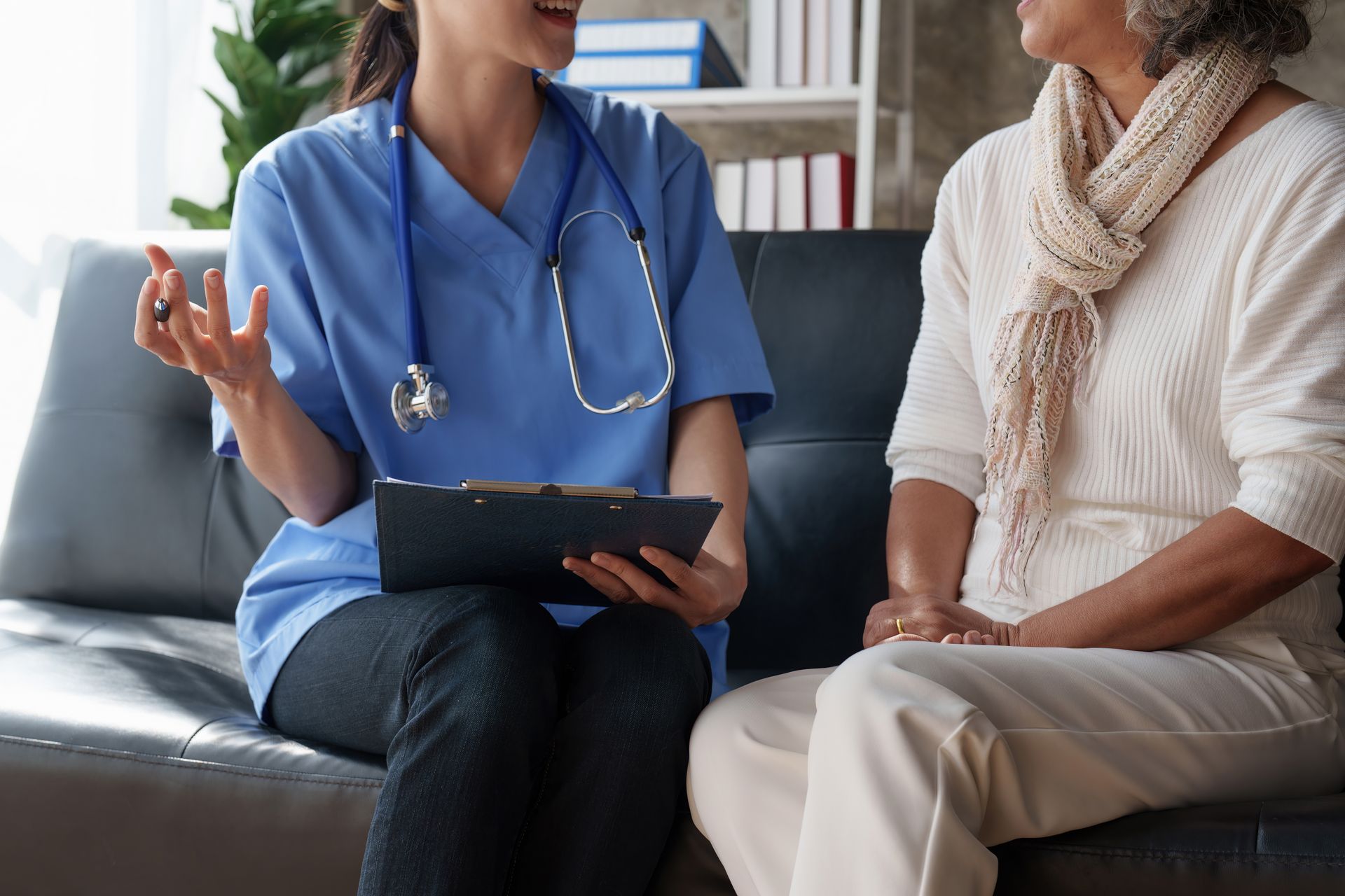 A medical professional in blue scrubs sits on a couch, holding a clipboard and talking with a patient wearing a scarf. A medical professional in blue scrubs sits on a couch, holding a clipboard and talking with a patient wearing a scarf.